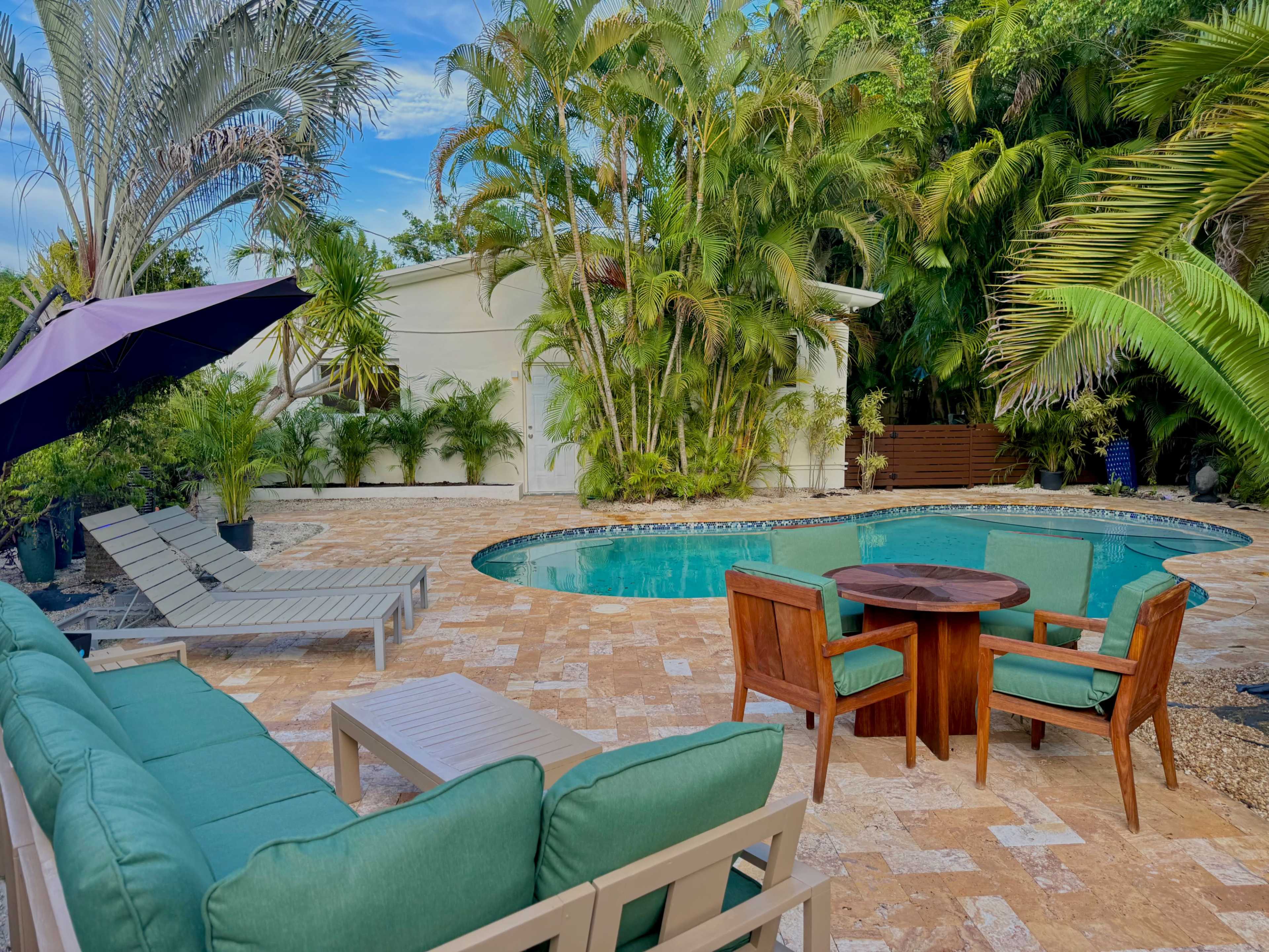 A spacious outdoor patio features a turquoise pool surrounded by lounge chairs, a dining table, and lush tropical plants.