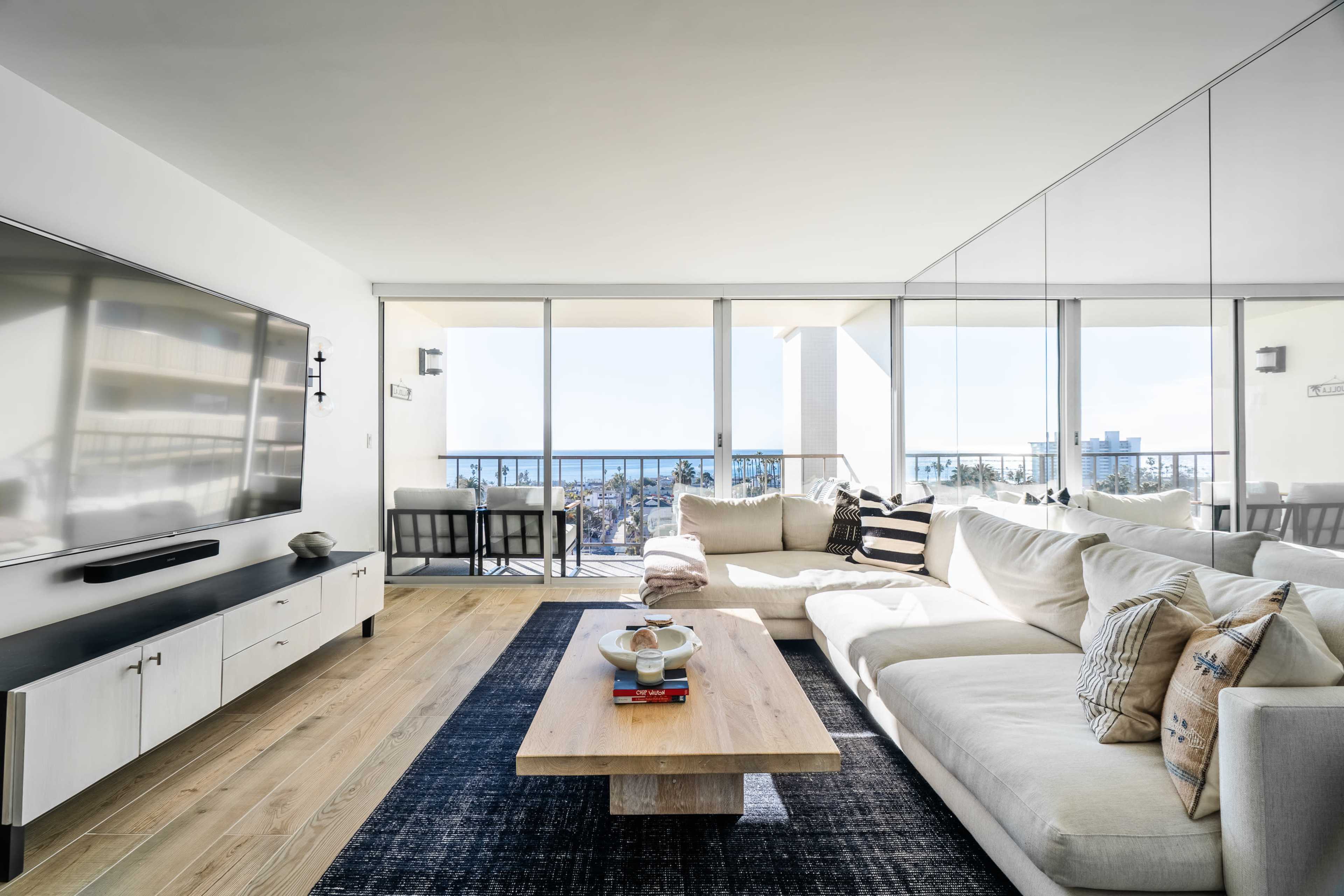 A modern living room features a large sectional sofa, a low wooden coffee table, and a wall-mounted television, with expansive glass doors offering a view of a city skyline.