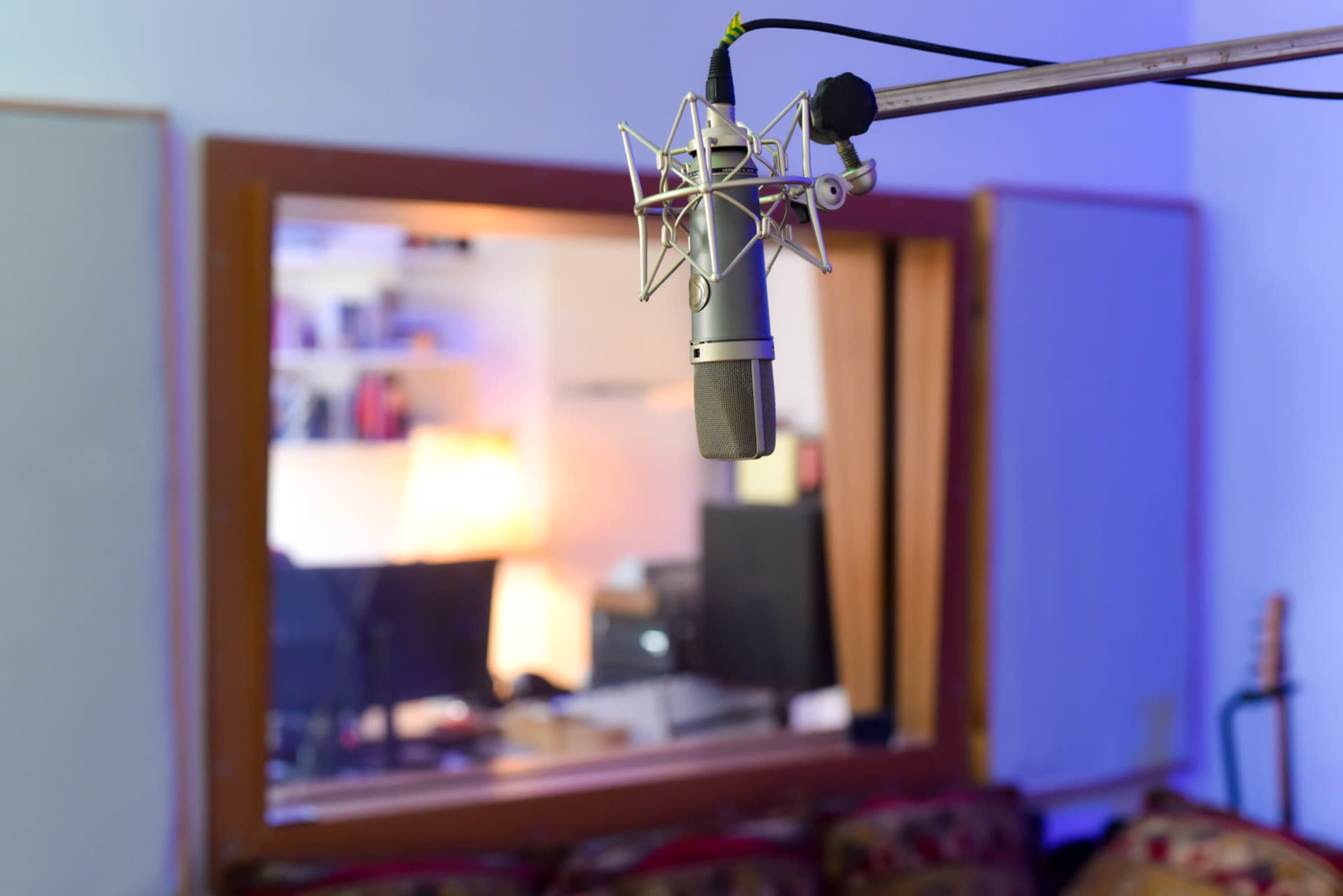 A microphone is positioned in front of a glass window revealing a recording studio with visible monitors and furniture.