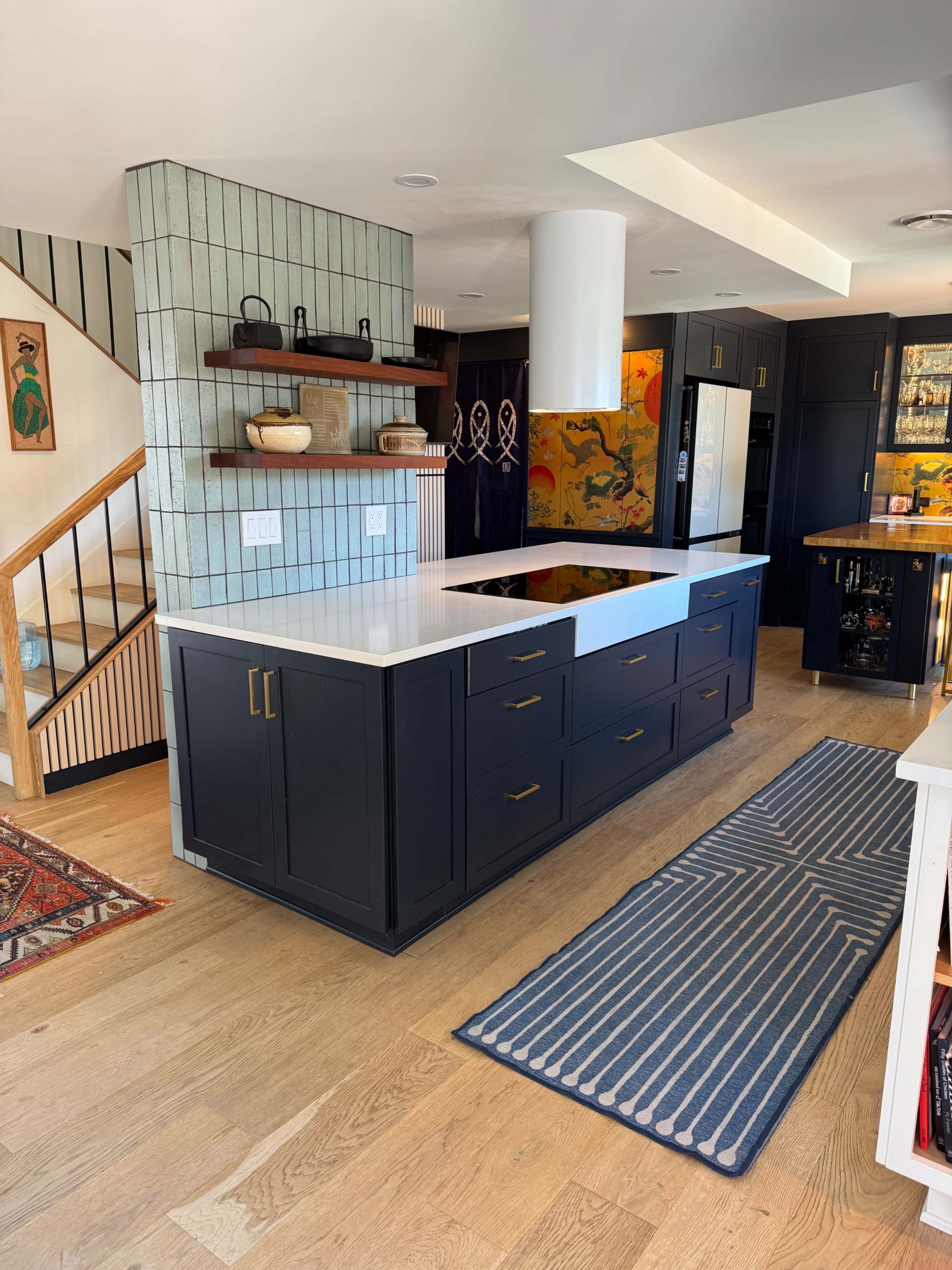 The image shows a modern kitchen featuring a central island with a white countertop, navy blue cabinets, a patterned backsplash, and a striped area rug on a wooden floor.