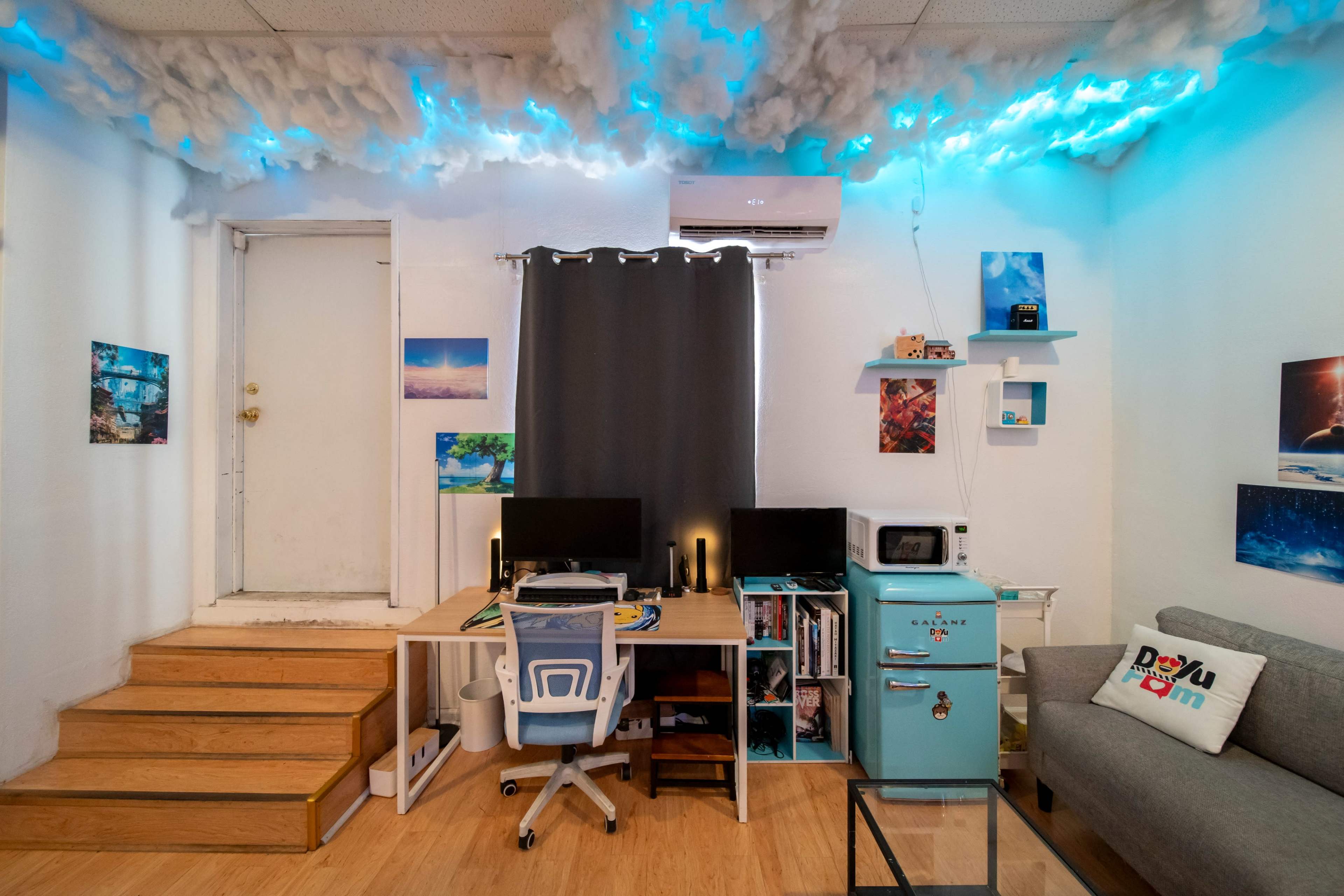 The image shows a small, modern workspace with two computer monitors on a desk, a retro-style refrigerator, and light blue cloud-like decorations on the ceiling.