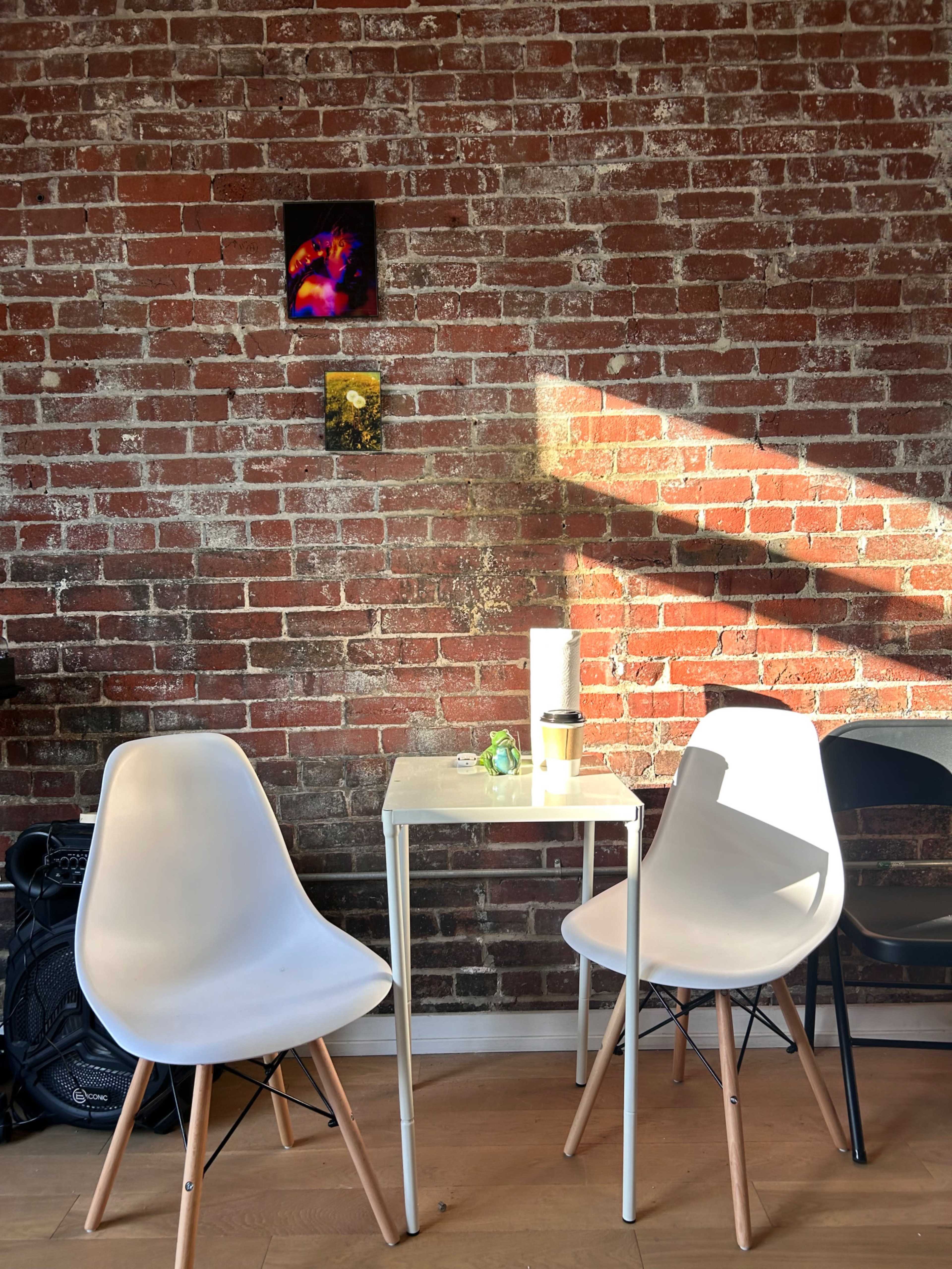 A small table with two chairs sits against a brick wall, illuminated by sunlight streaming through a nearby window, with artwork hanging above.
