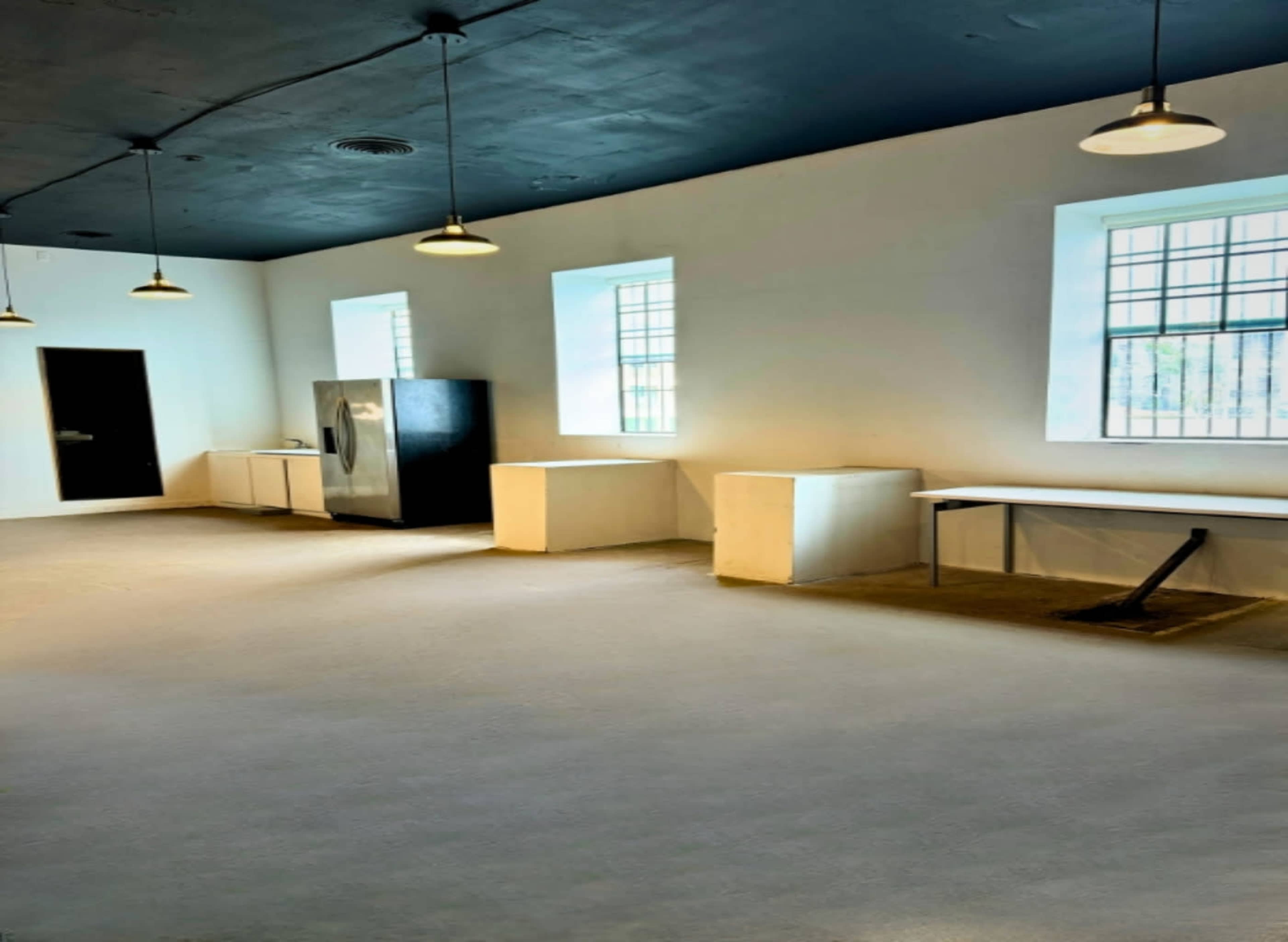 The image shows a spacious, minimally decorated room with white walls, a black ceiling, a stainless steel refrigerator, and a worktable, illuminated by pendant lights.