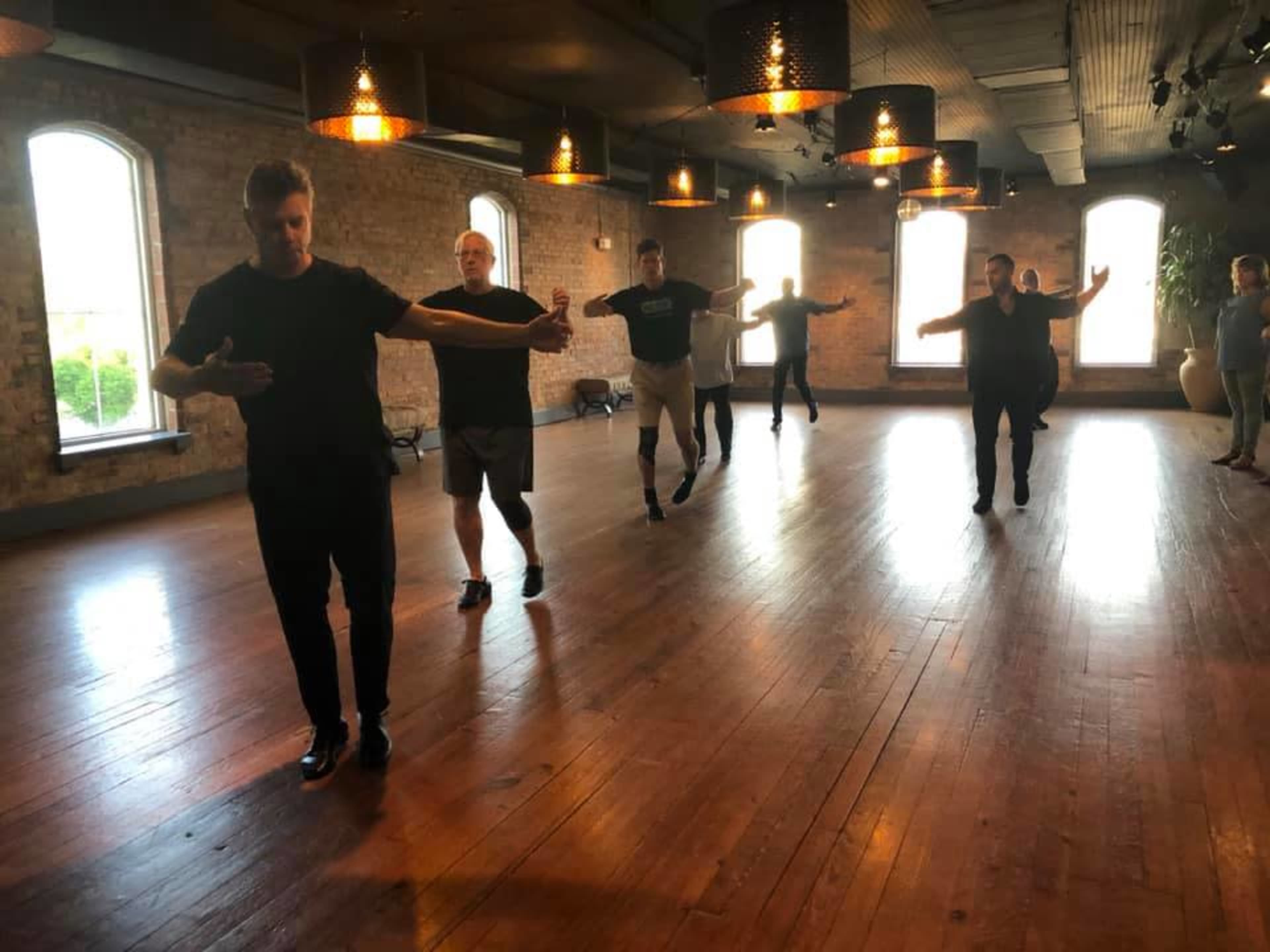 A group of six men are practicing dance moves in a well-lit, wooden-floored studio with exposed brick walls.