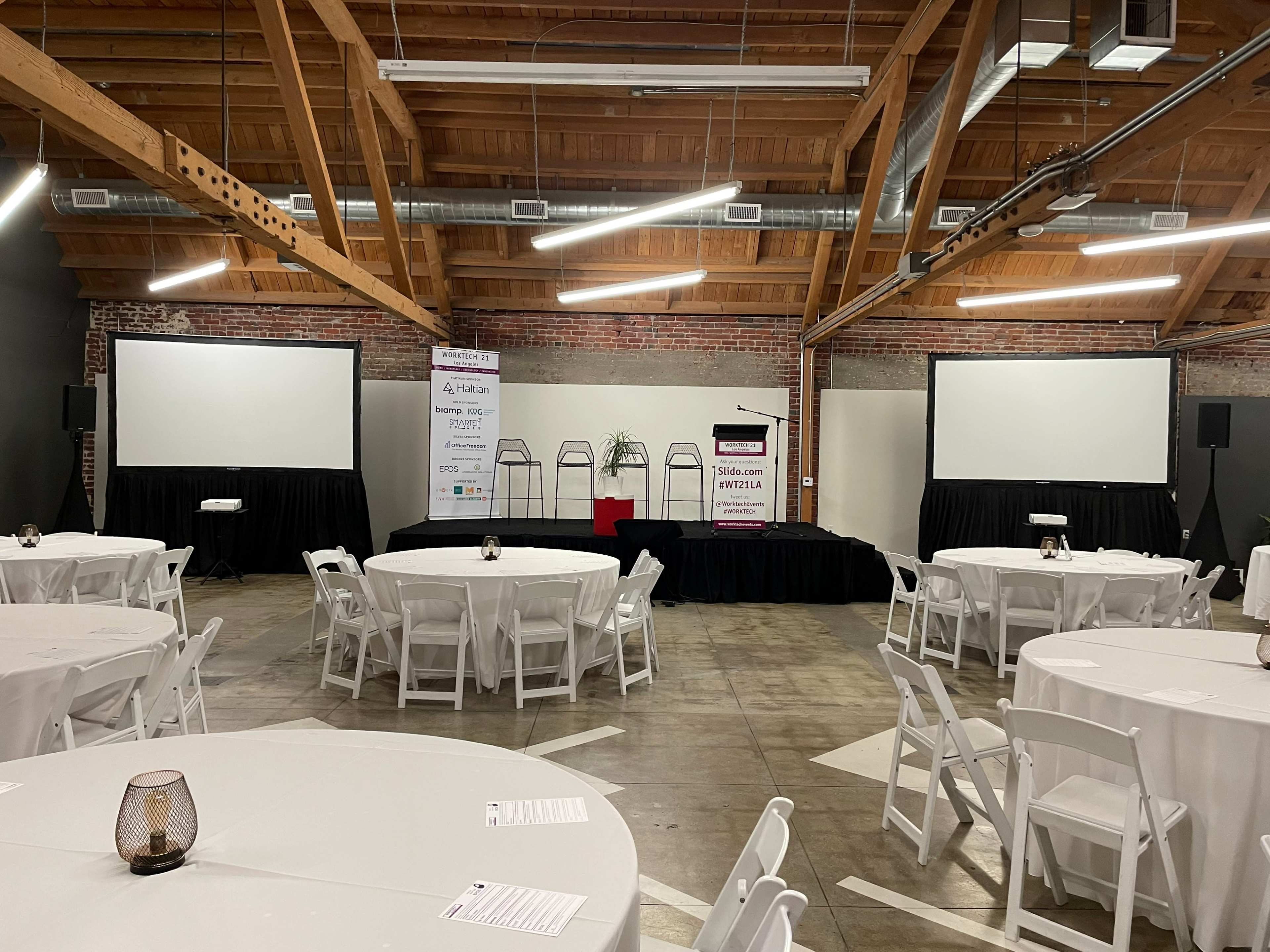 A spacious event venue set up with round tables, a stage, and large screens in a rustic industrial space.