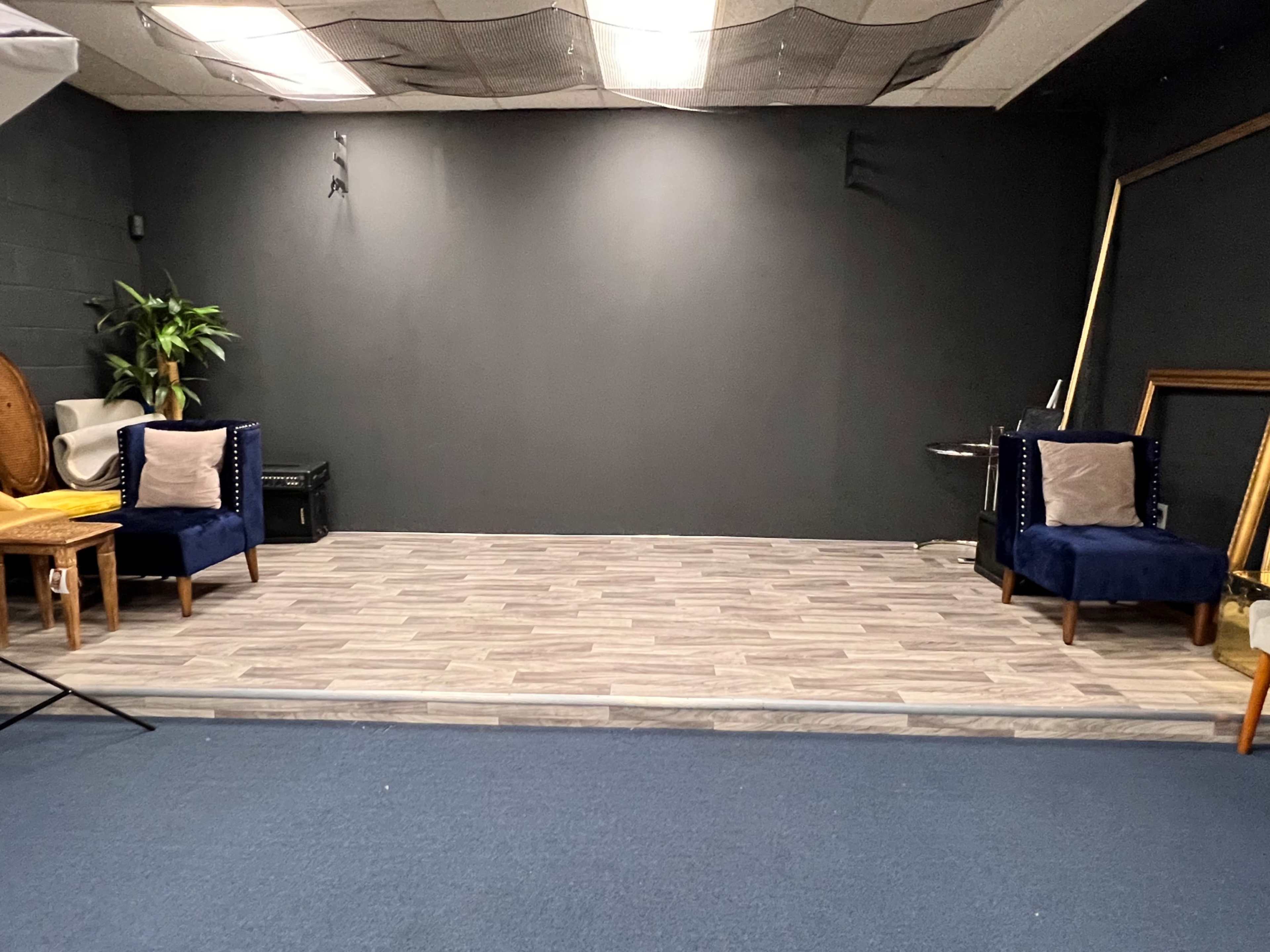 A stage area with two blue chairs, a small table, and a dark wall in the background.