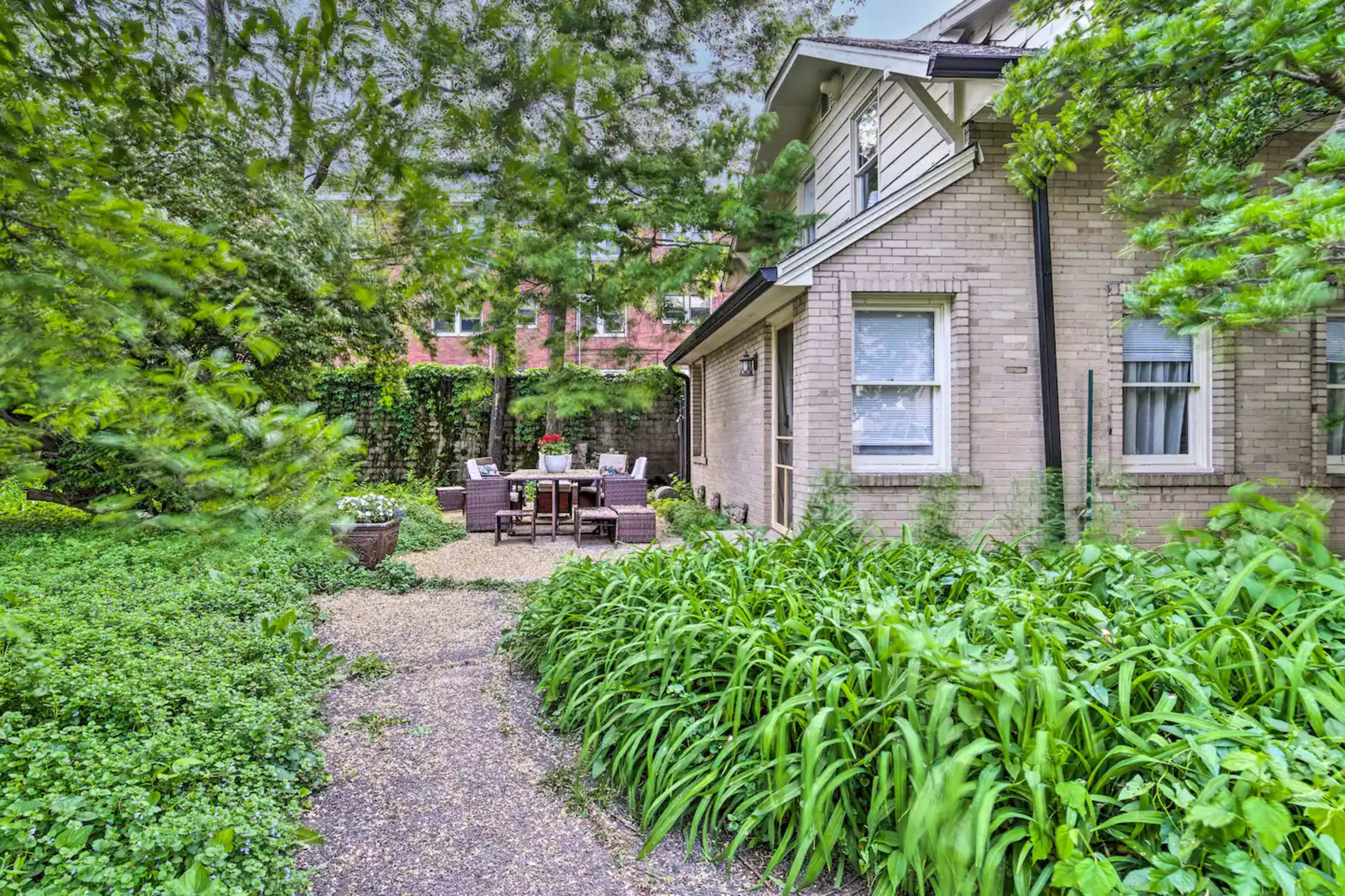 A pathway lined with greenery leads to a seating area with furniture set up outside a brick house.