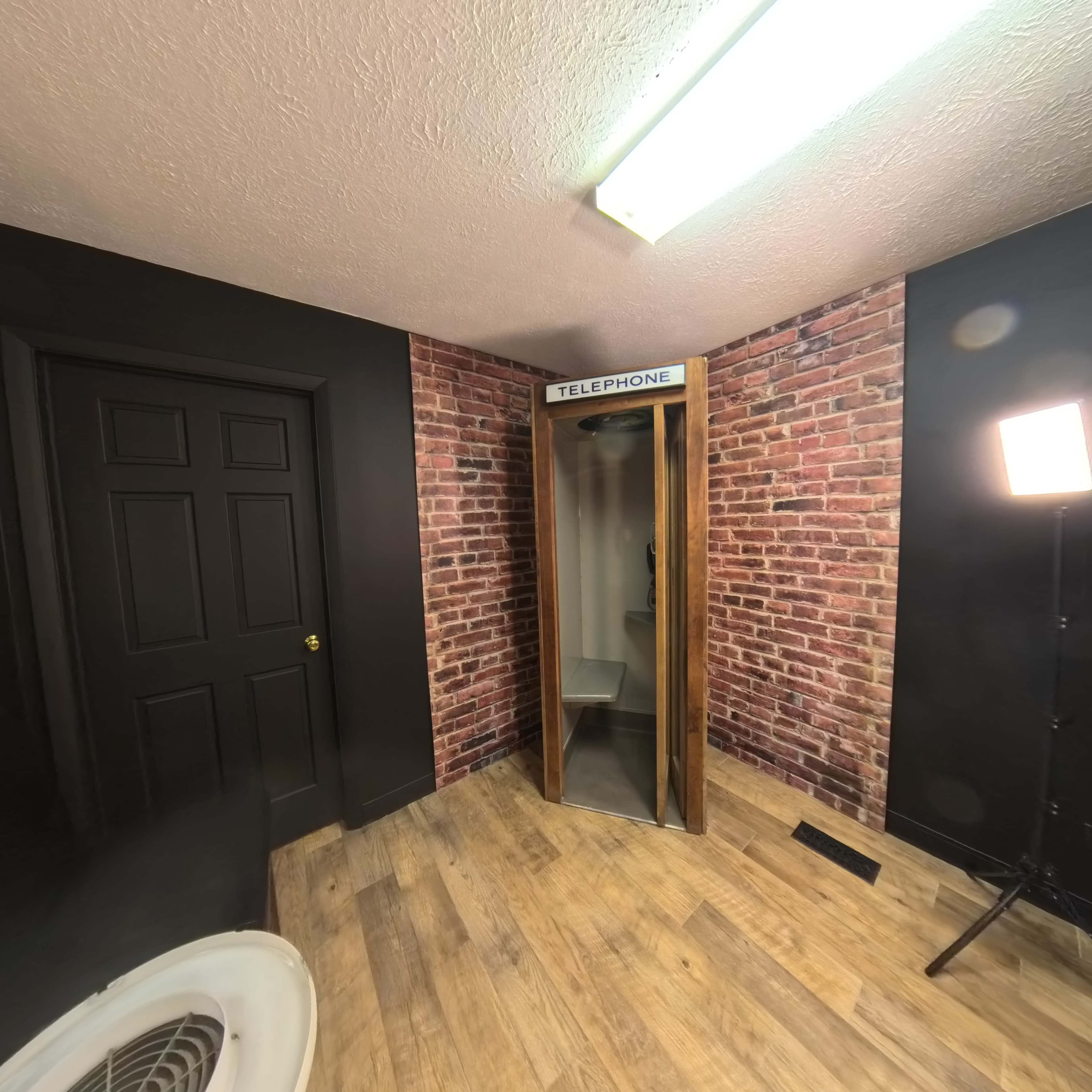 The image shows a vintage telephone booth situated in a room with exposed brick walls and a wooden floor.