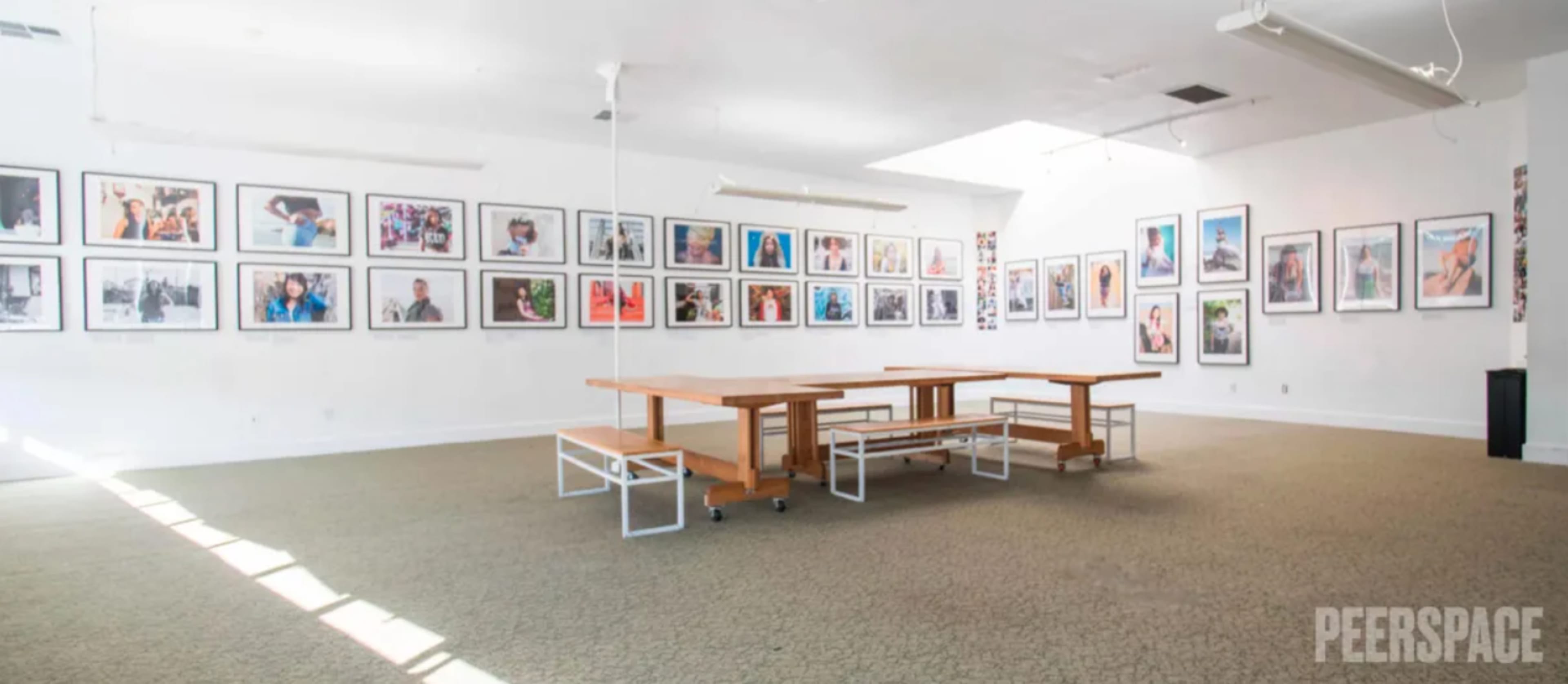 The image shows a spacious gallery with white walls adorned with framed photographs, and a wooden table with benches set in the center.
