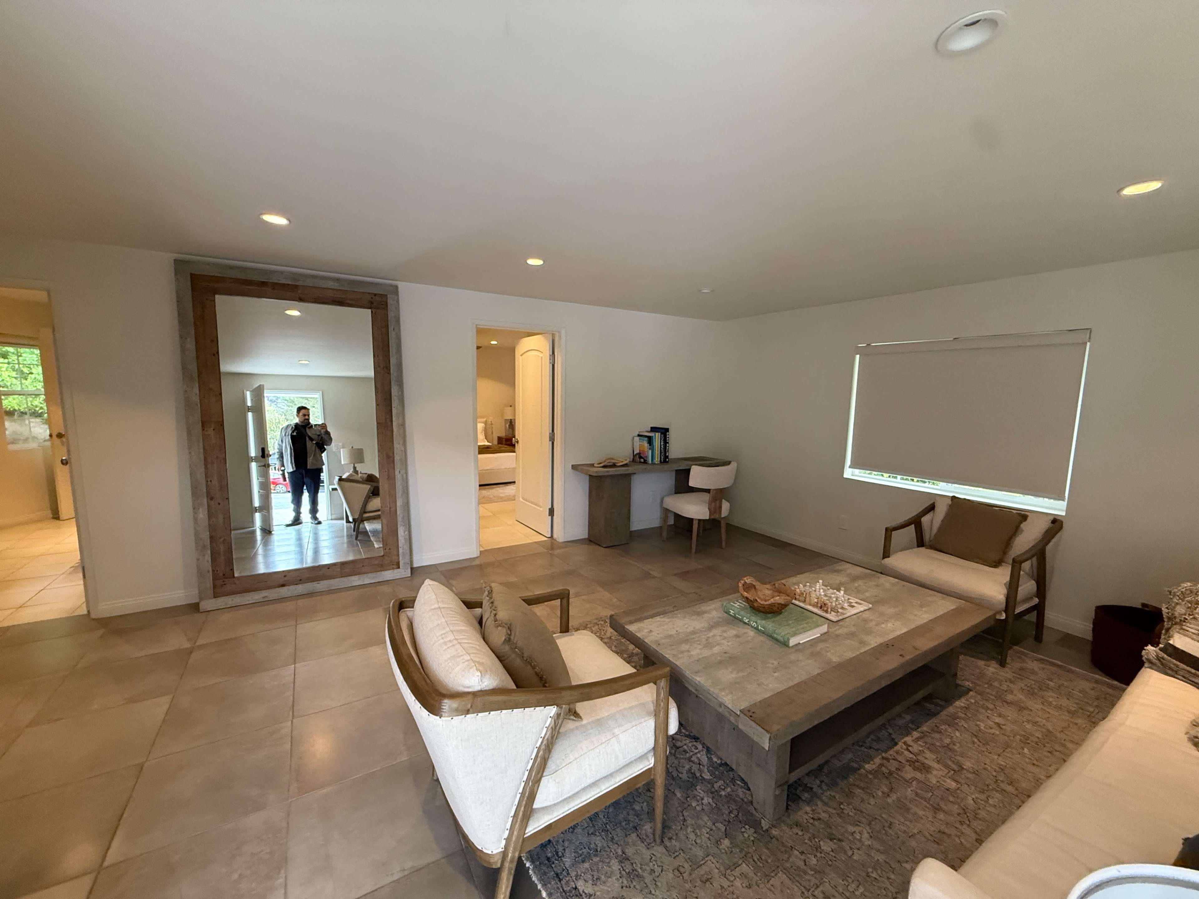 The image shows a spacious living room with a central table, two armchairs, a large mirror on the wall, and a doorway leading to another room.