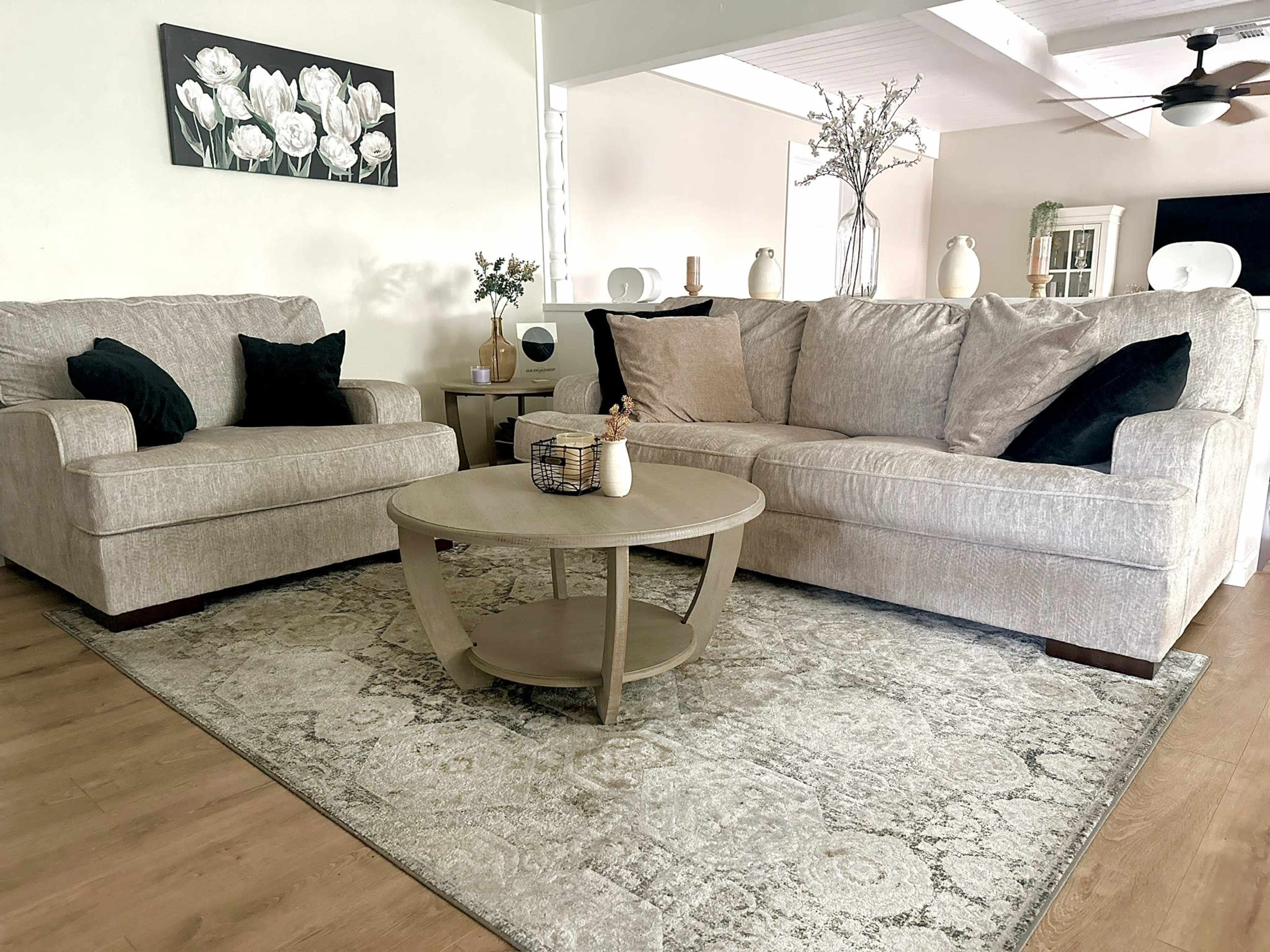 A living room features two light-colored sofas with black throw pillows arranged around a round coffee table on a patterned rug.