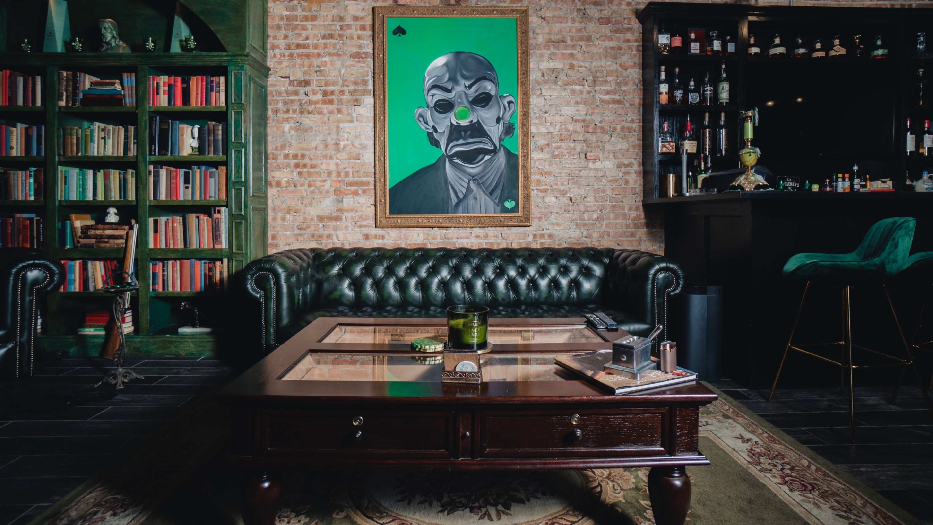 A leather tufted sofa faces a wooden coffee table on a patterned rug, with a colorful portrait on the brick wall above and bookshelves to the left.