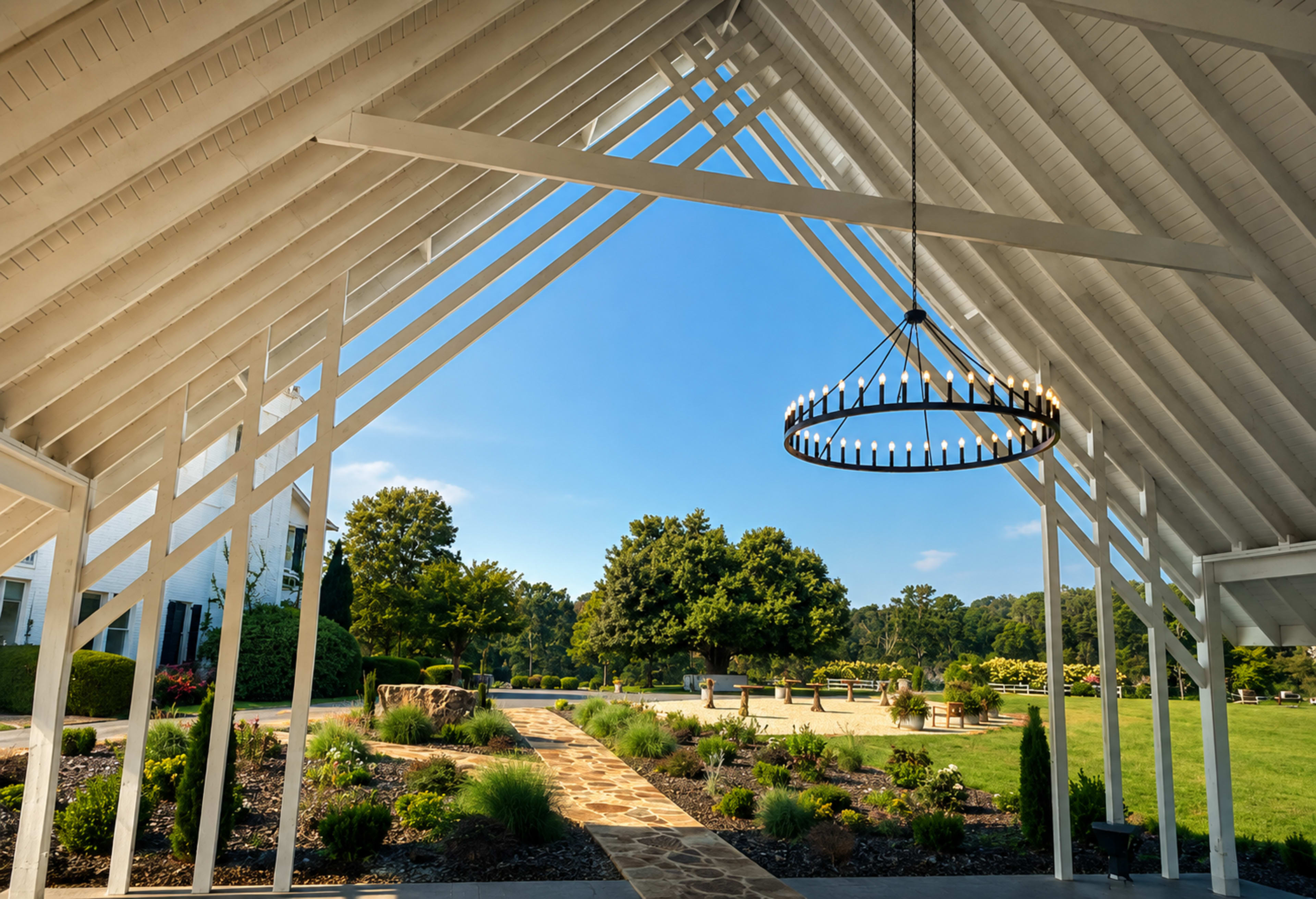 The image shows a spacious outdoor pavilion with a white wooden frame, featuring a circular chandelier, overlooking a landscaped garden path and lush greenery.
