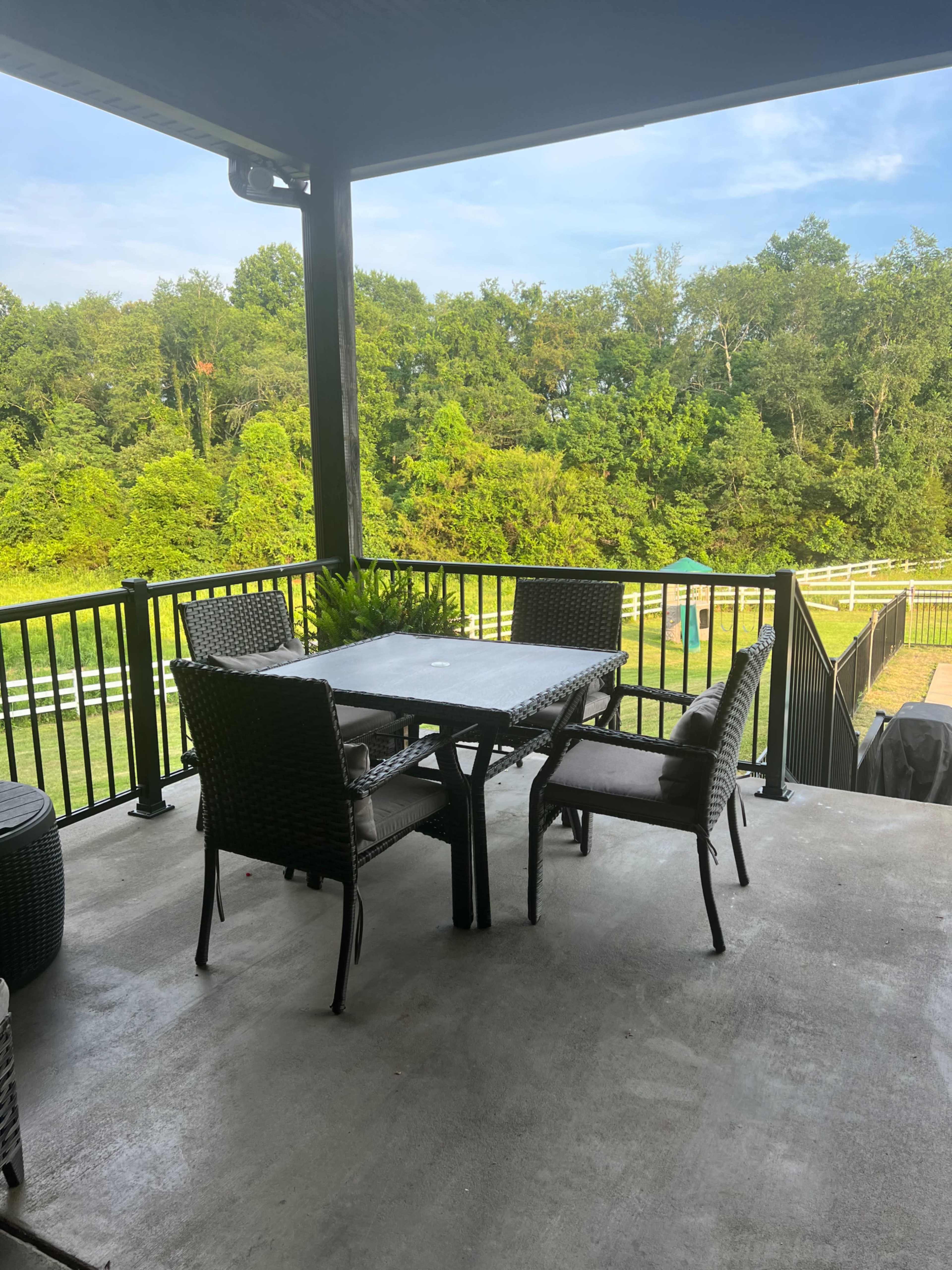 A patio with a table and chairs overlooks a green, wooded area and a fence.
