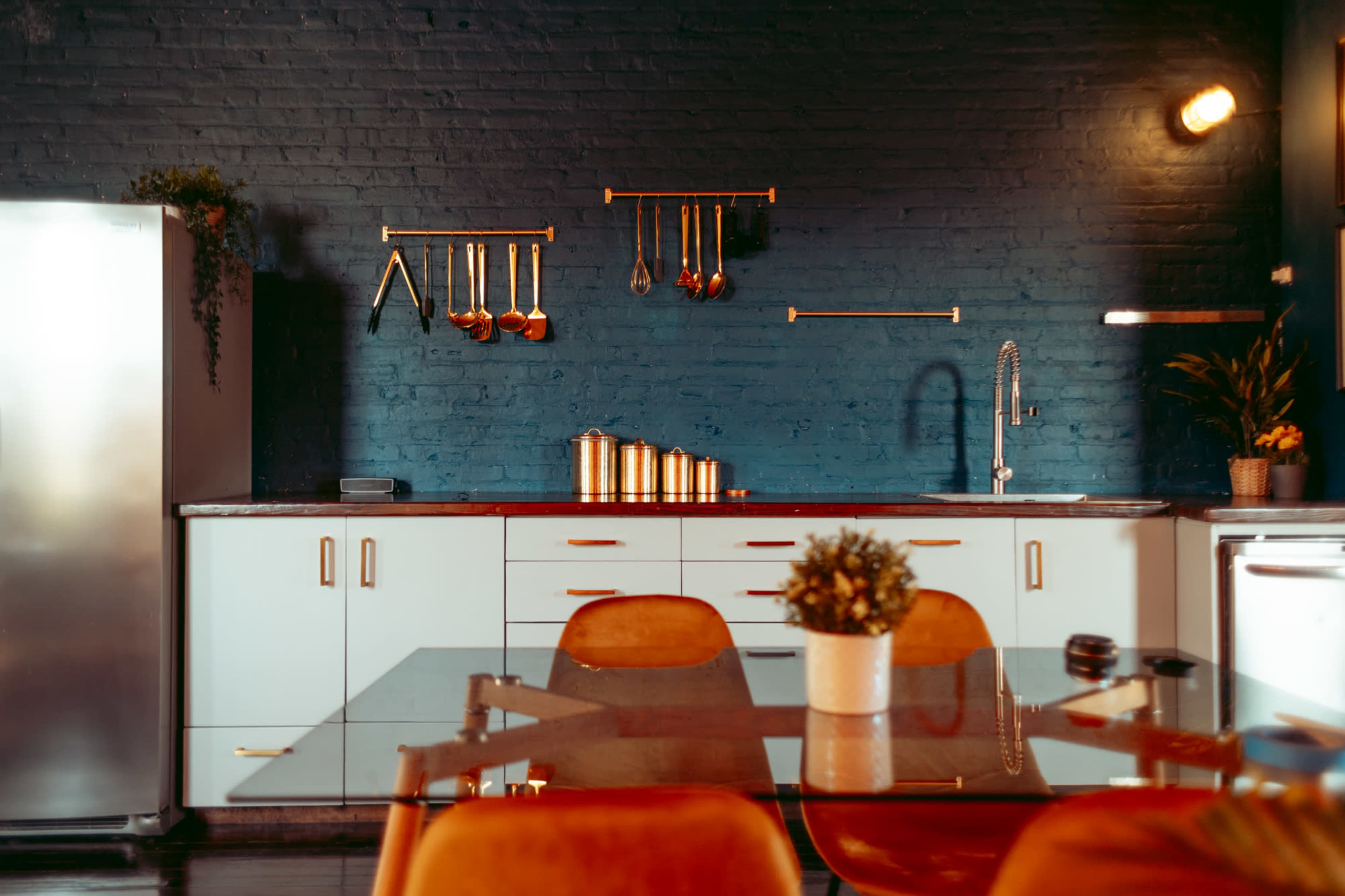 A modern kitchen features a dark brick wall, stainless steel appliances, and a glass-top dining table surrounded by orange chairs.