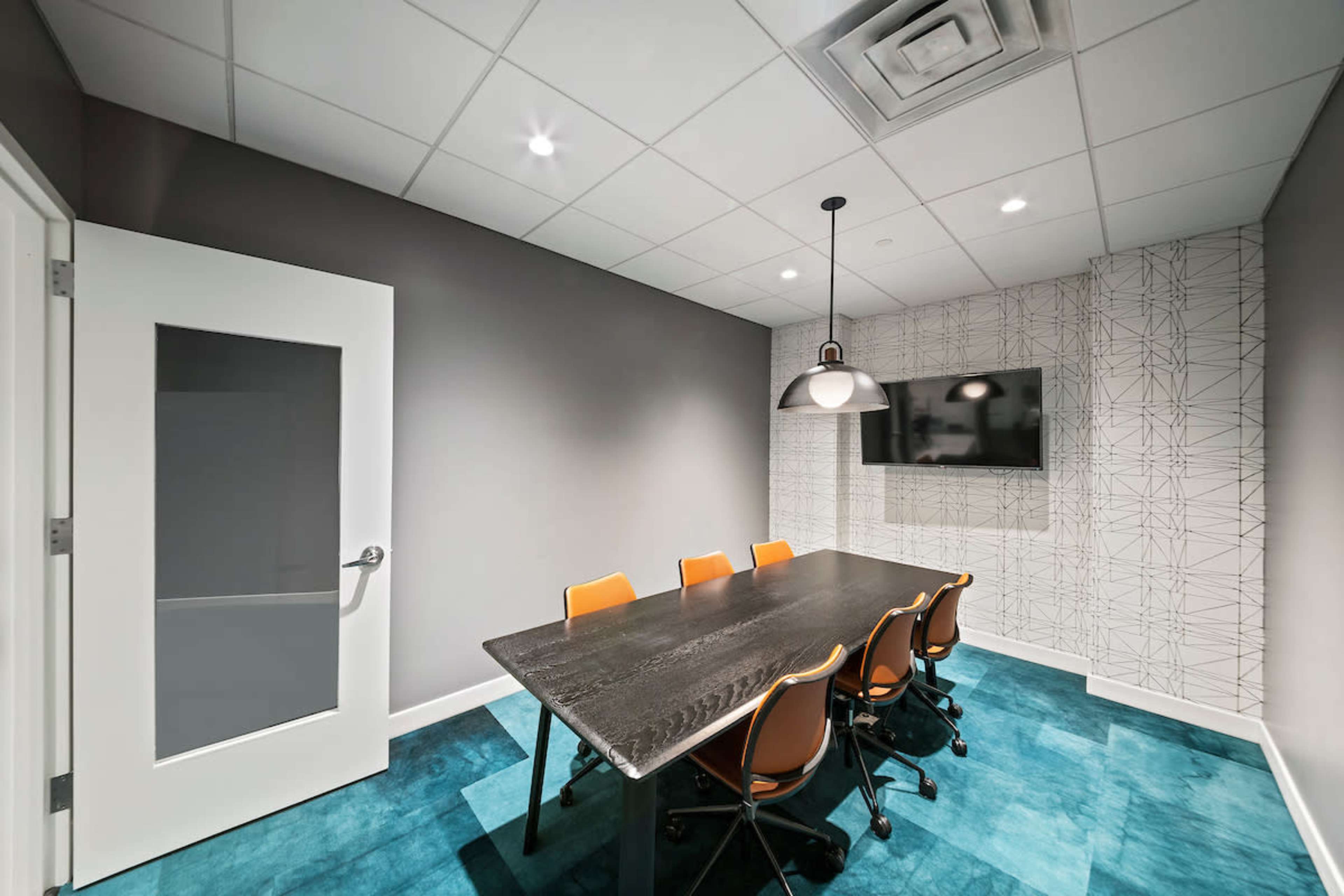 A conference room features a large black table surrounded by orange chairs, with a television on the wall and a patterned accent wall.