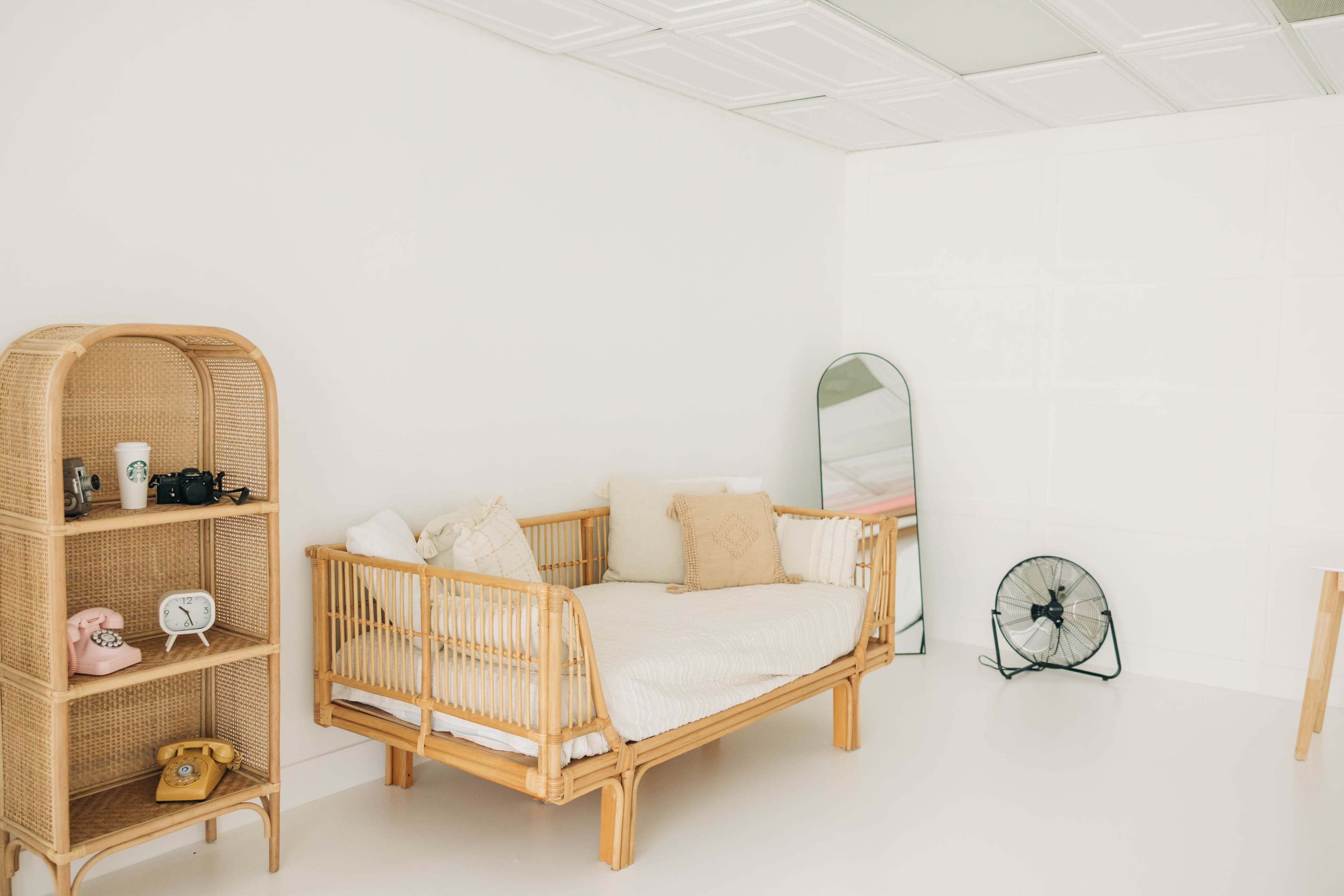 The image shows a bright, minimalist room featuring a rattan daybed, a nearby shelf with vintage cameras, and a large mirror against the white wall.