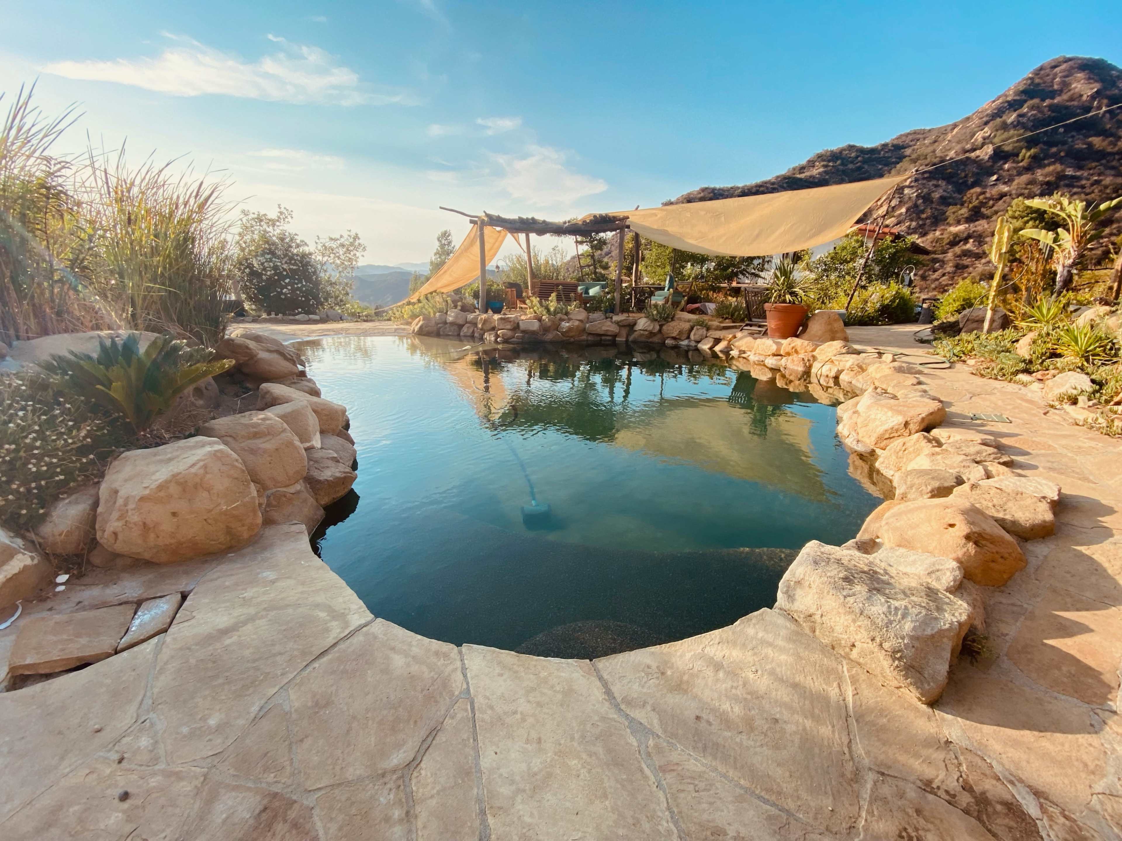 A natural stone pool surrounded by lush vegetation and a shaded seating area, set against a backdrop of rolling hills.