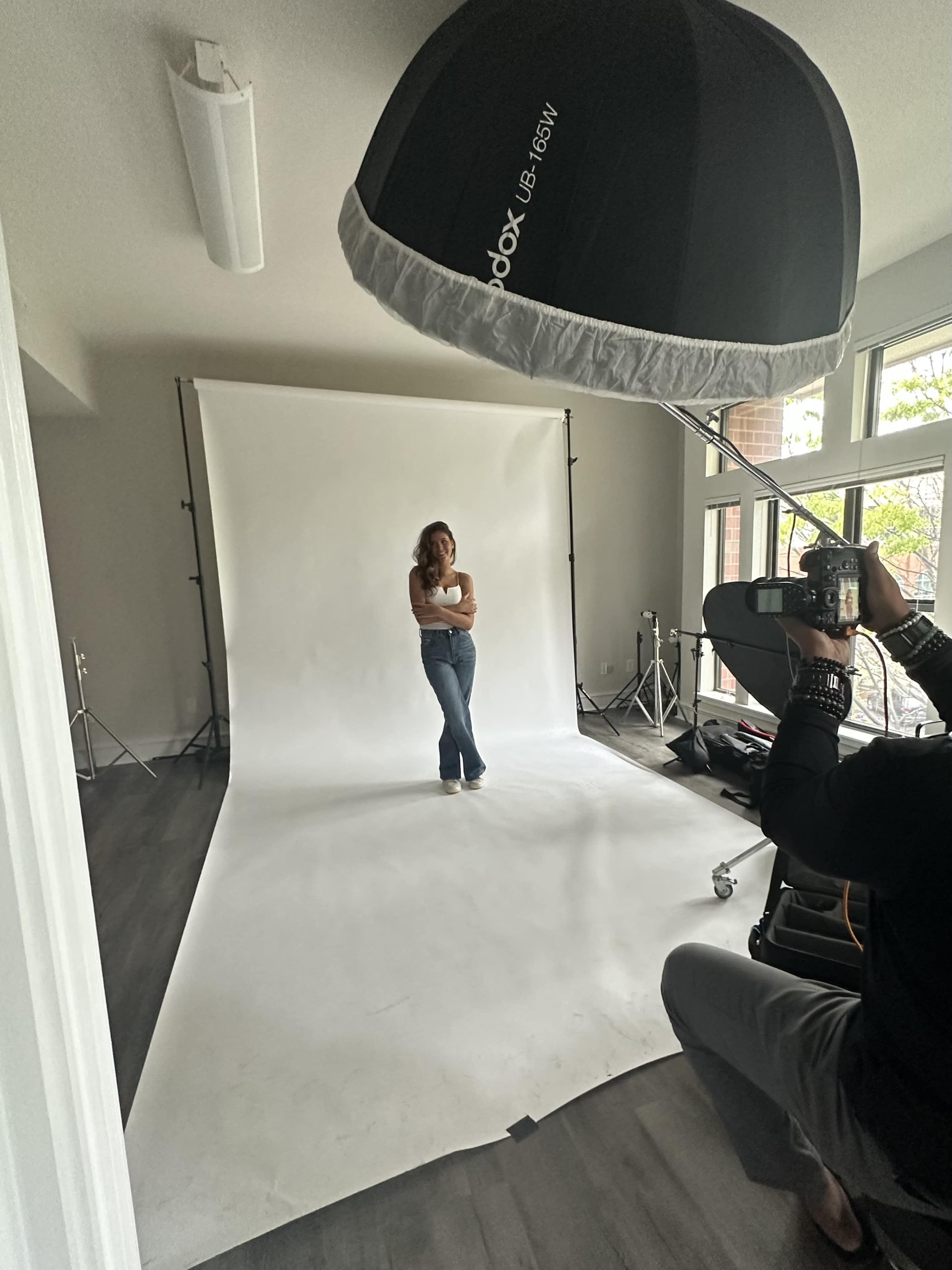 A woman posing in front of a white backdrop is illuminated by a large studio light while a photographer captures the moment.