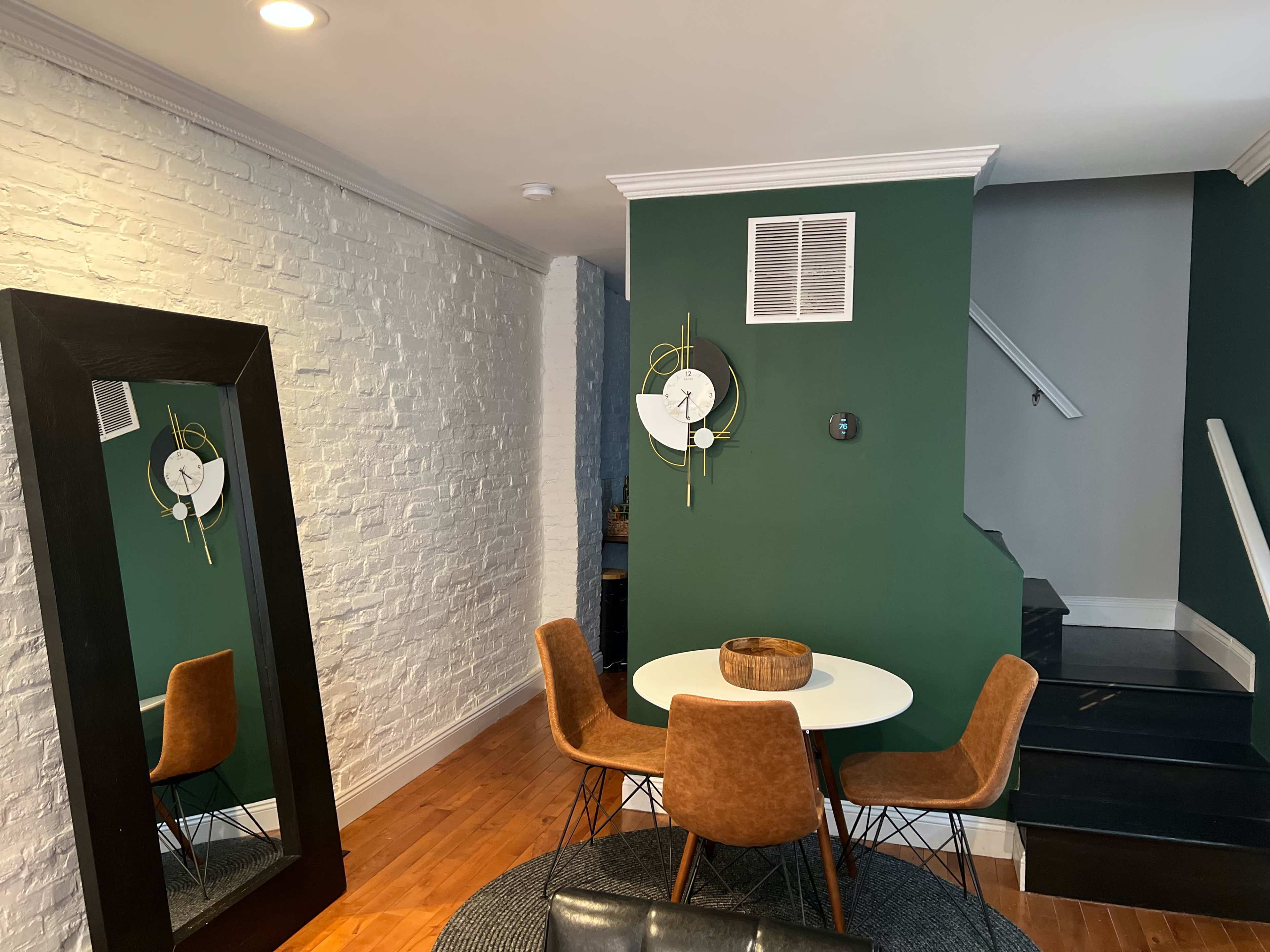 The scene features a modern dining area with a round white table surrounded by four brown chairs, a large mirror on the left, and a dark staircase in the background.