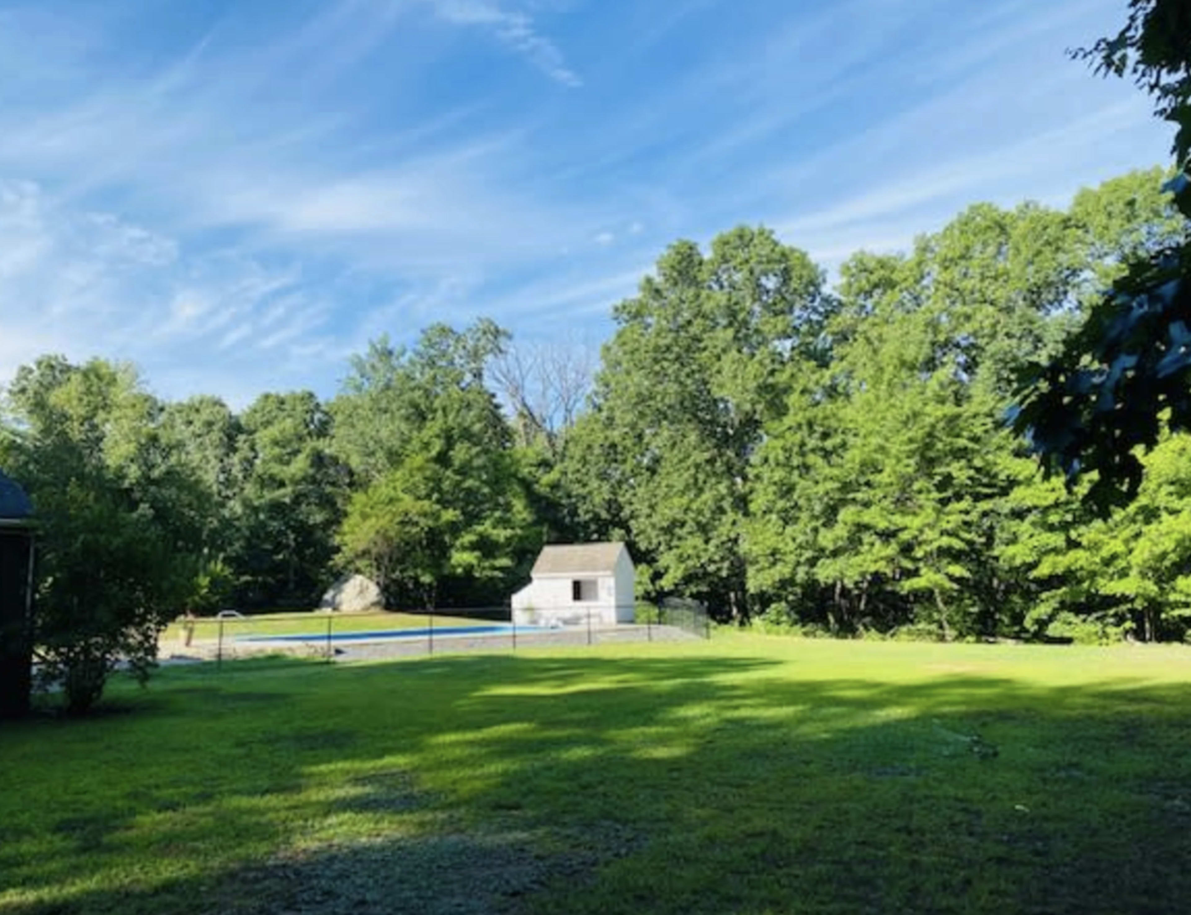 The image shows a grassy field with a small white shed set against a backdrop of trees and a blue sky.