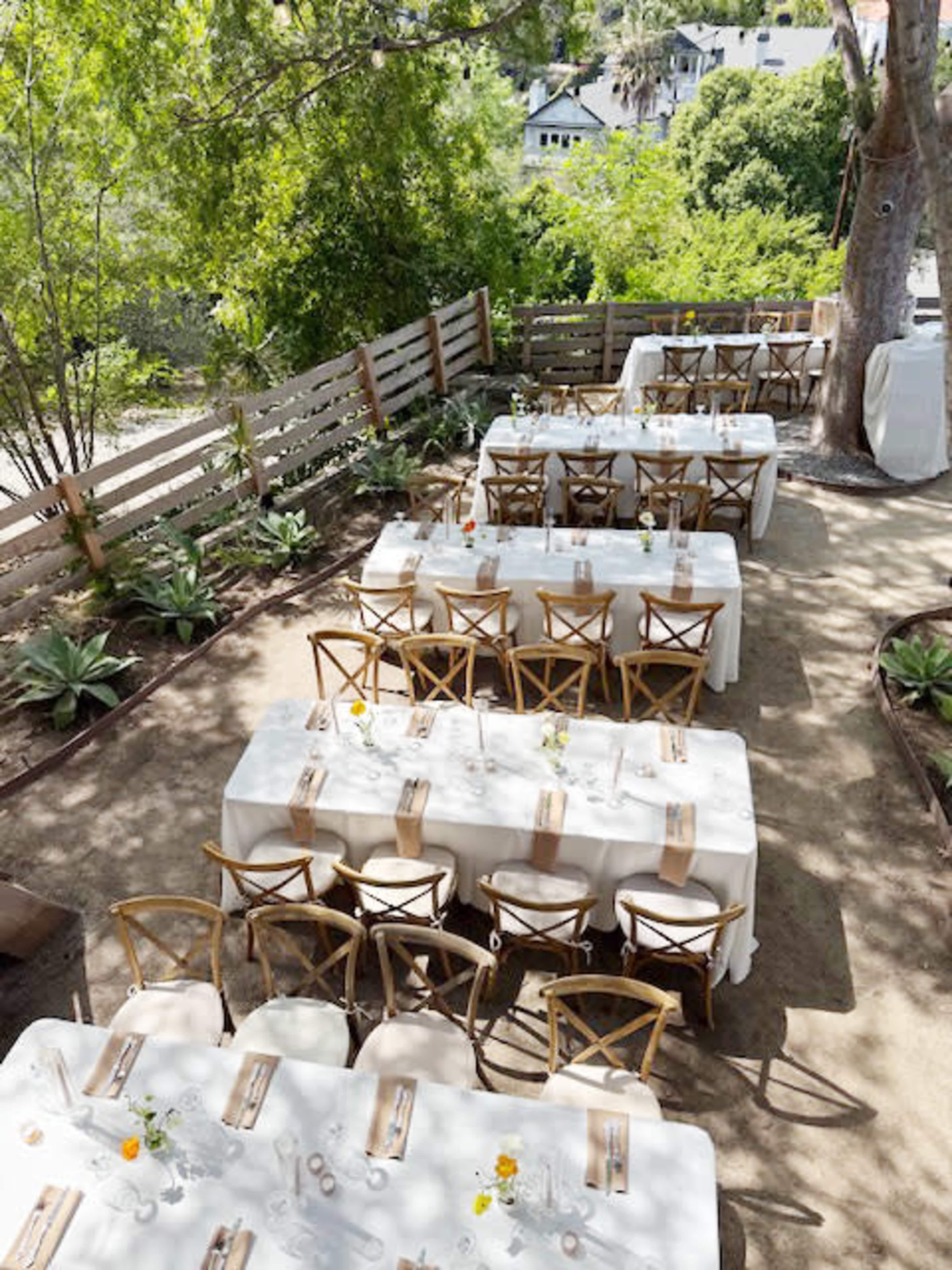 The image shows a wedding reception setup with multiple long tables covered in white tablecloths, arranged in an outdoor garden surrounded by greenery.