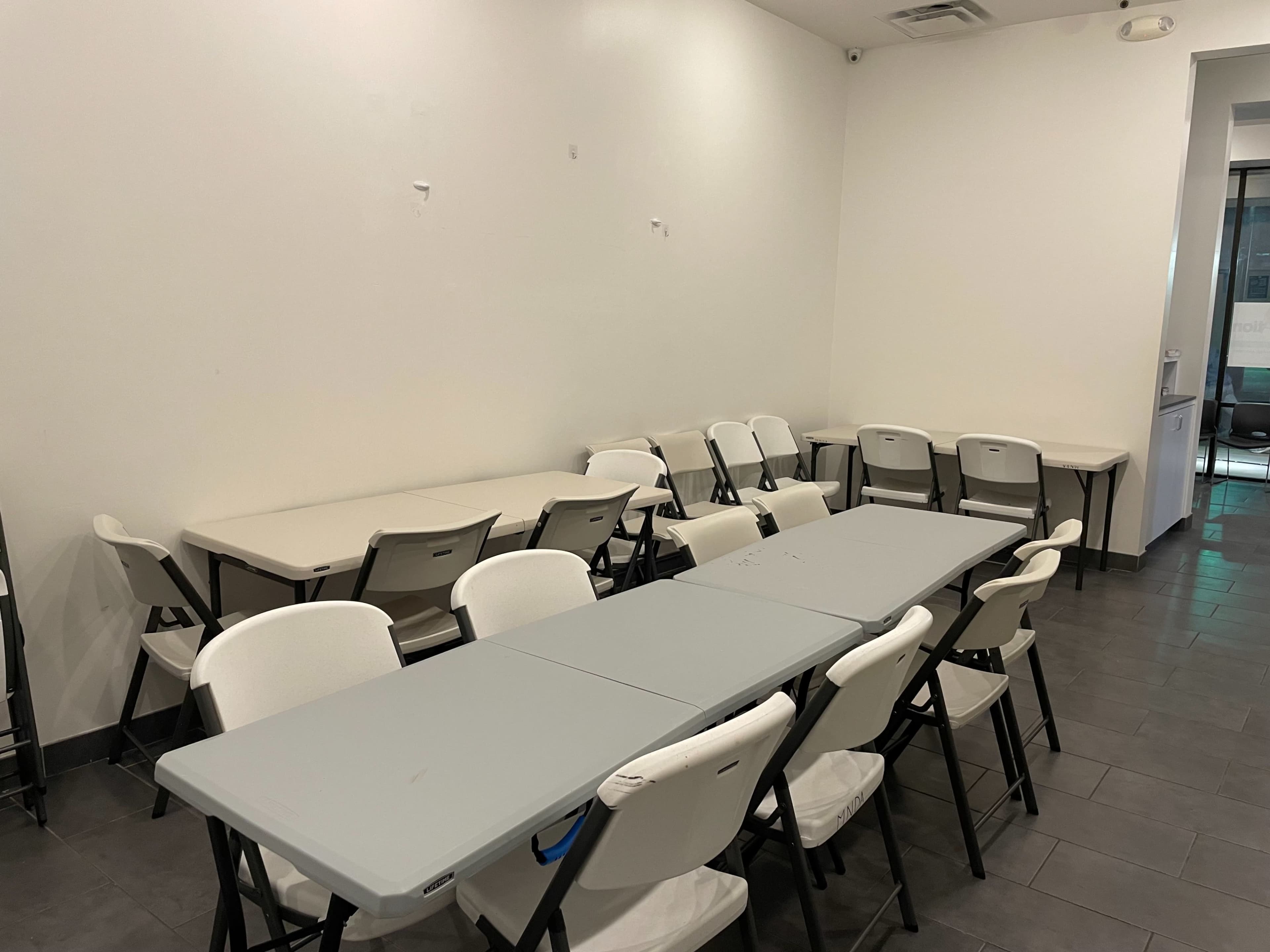 The image shows a small, empty room with several folding tables arranged in a U-shape and white folding chairs positioned around them.