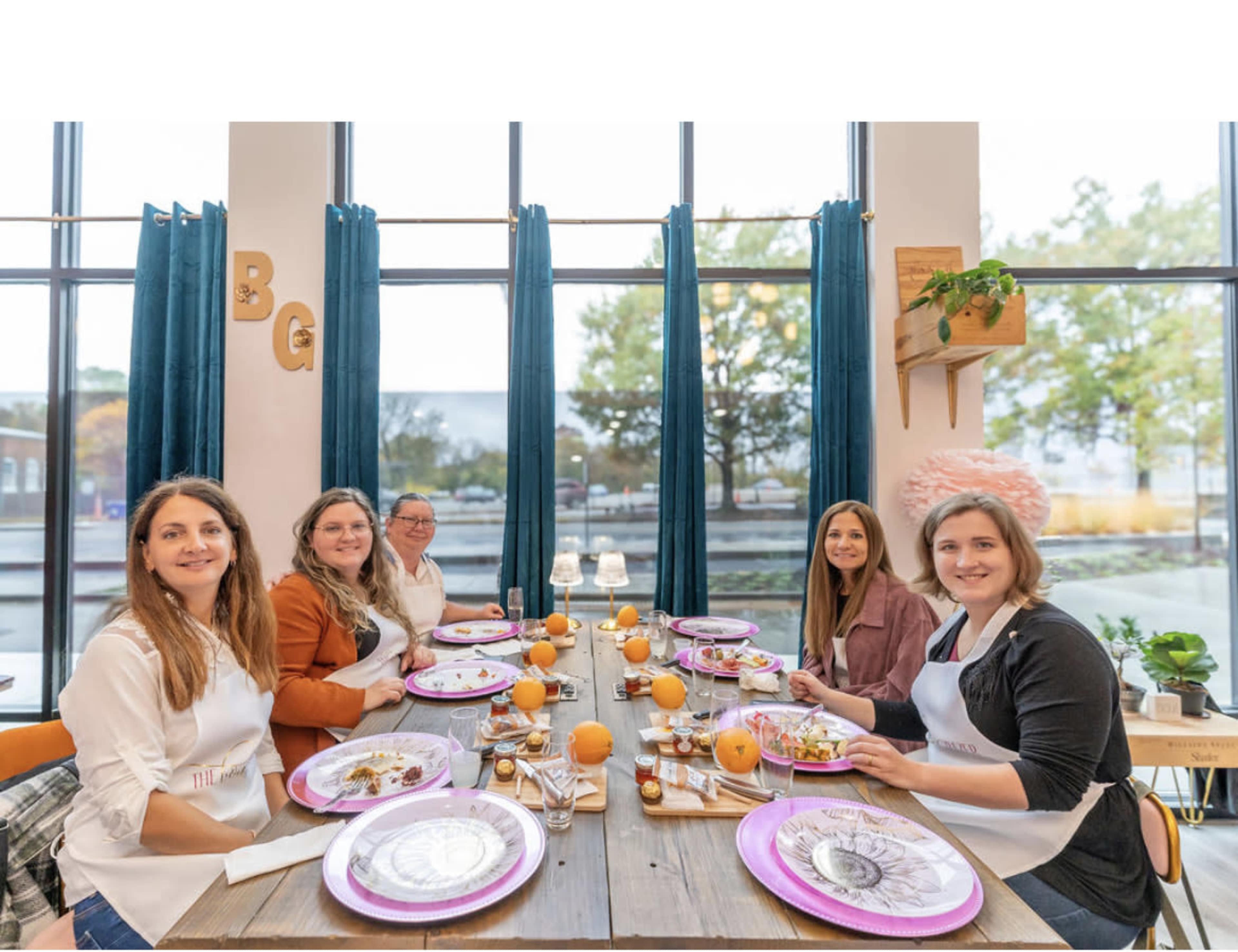 A group of five women sits at a long wooden table set with plates and food, surrounded by large windows and decorative elements.