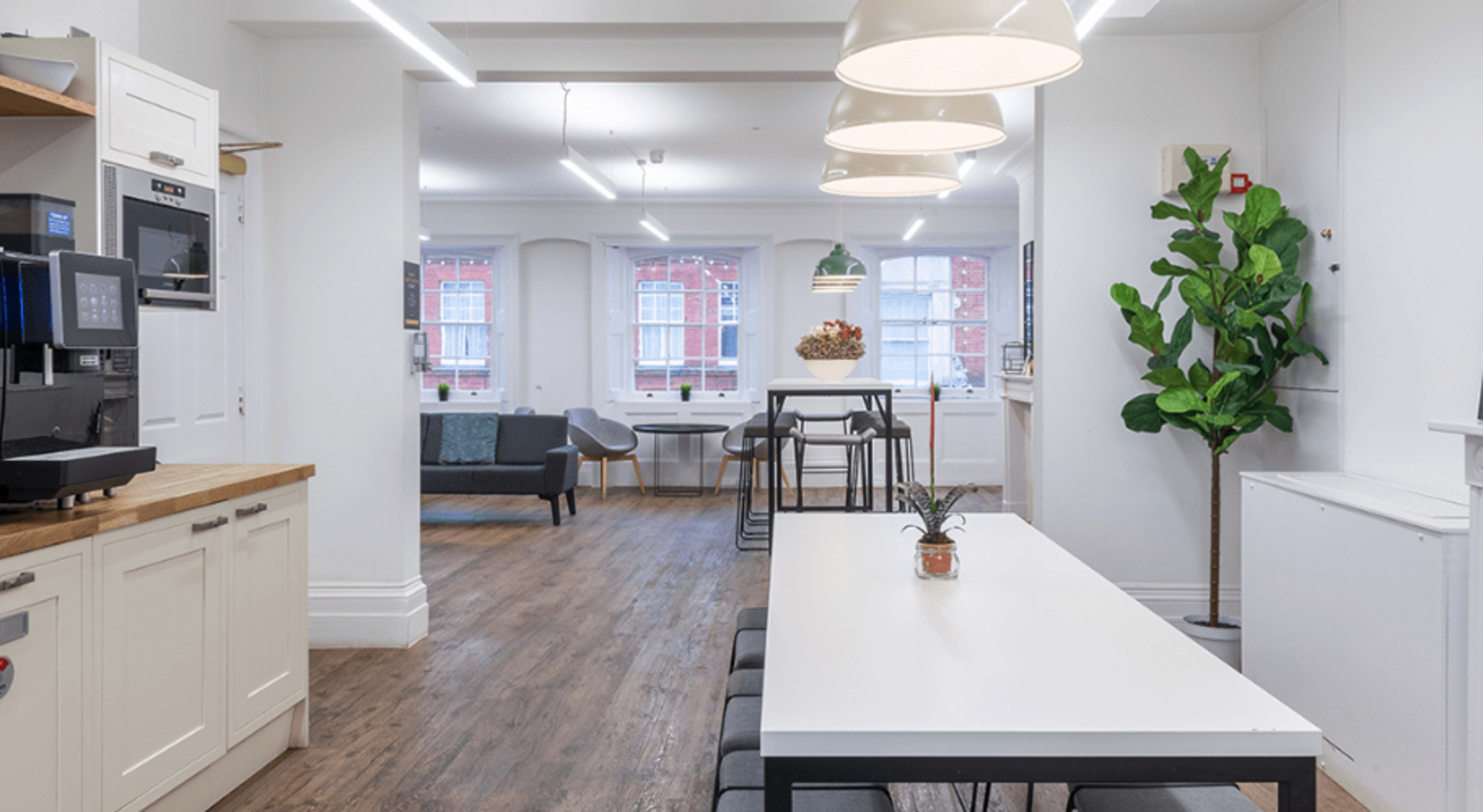 A modern kitchen and dining area with a long white table, chairs, a coffee machine, and a green plant, illuminated by pendant lights.