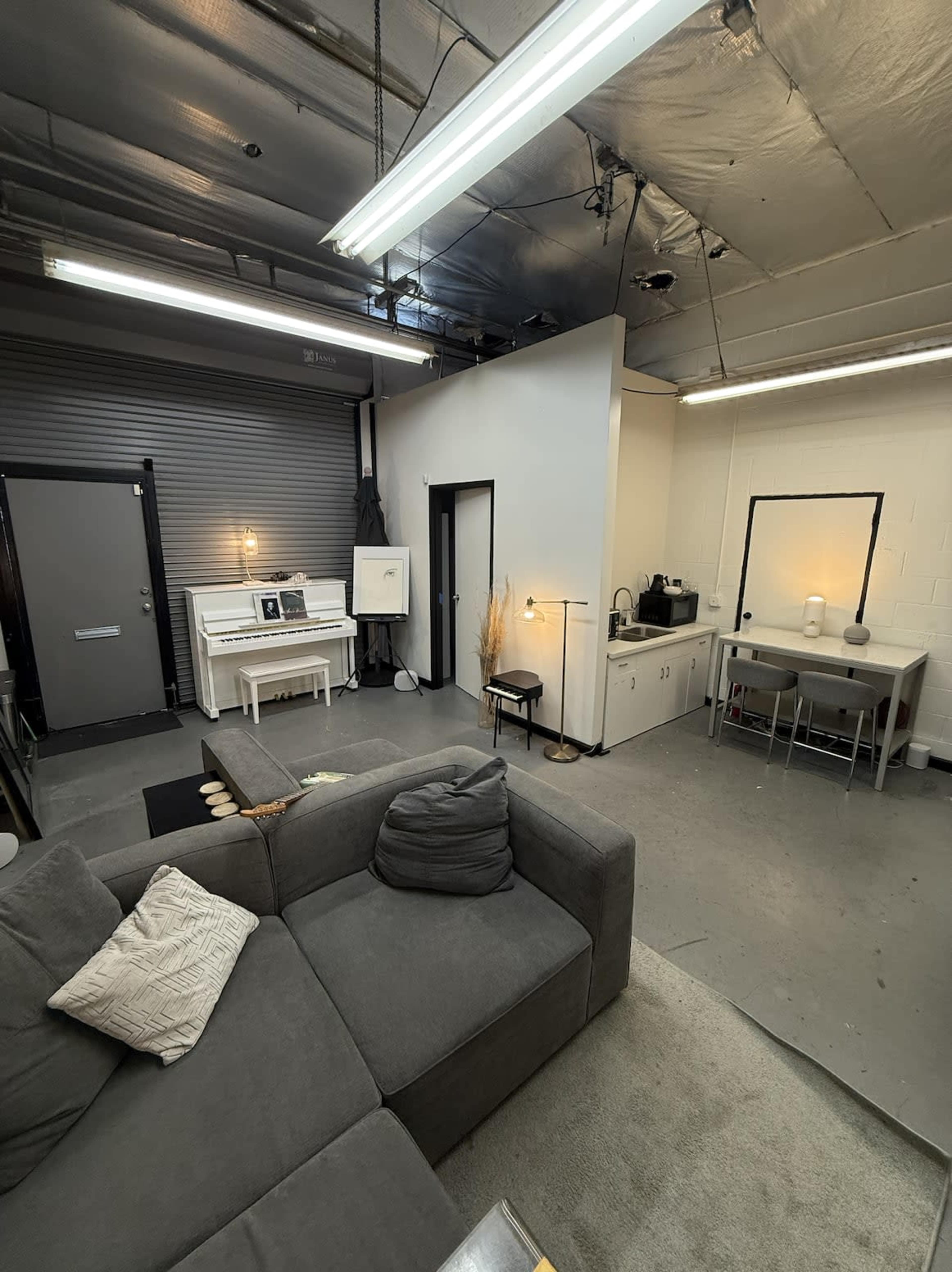 The image shows a spacious interior with a gray sectional sofa, a white piano, a small kitchen area, and a workspace with a table and chairs, all under a ceiling with exposed pipes and industrial lighting.