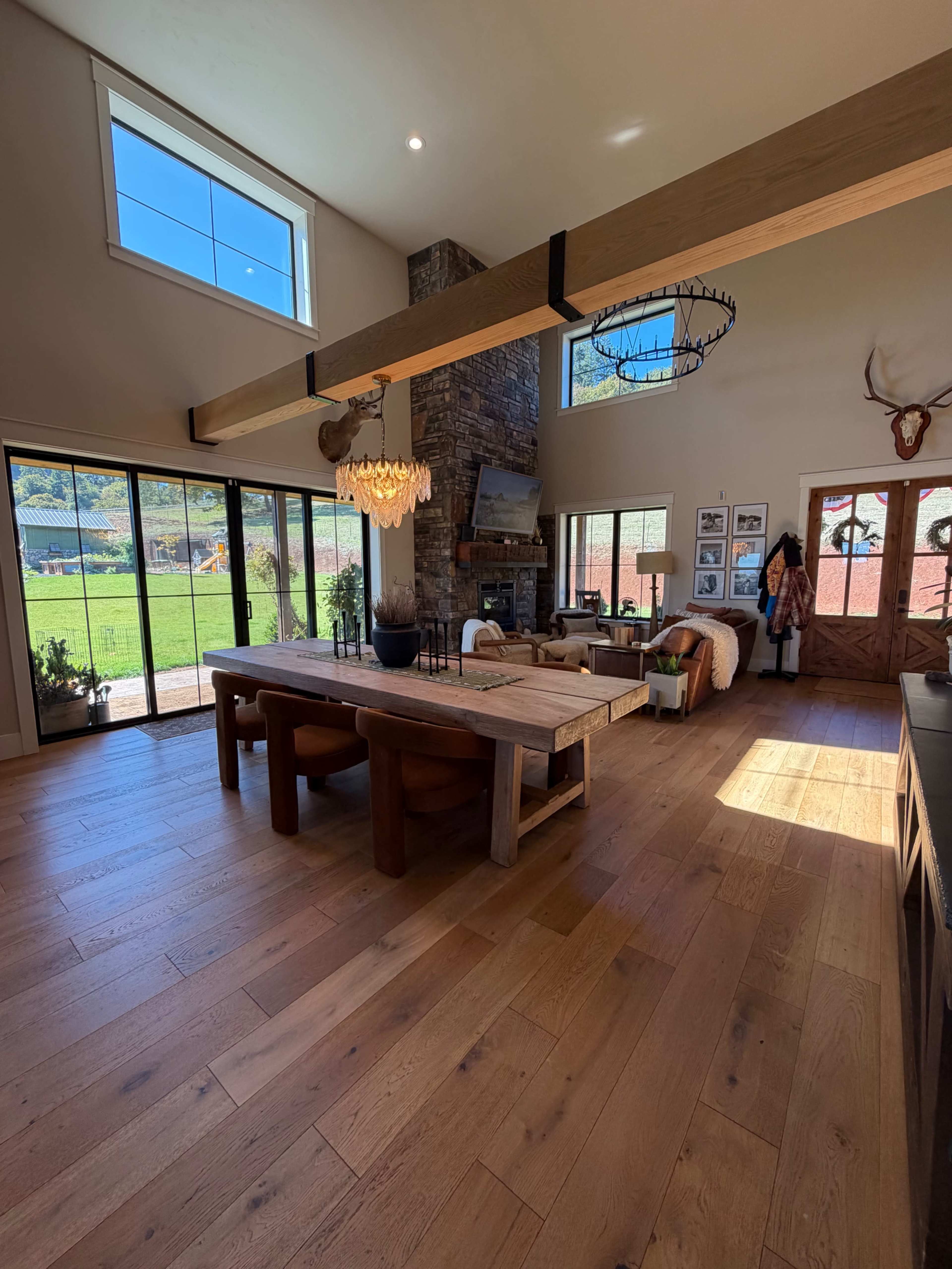 The image shows a spacious interior with a large wooden dining table, a stone fireplace, and large windows offering a view of the outdoors.