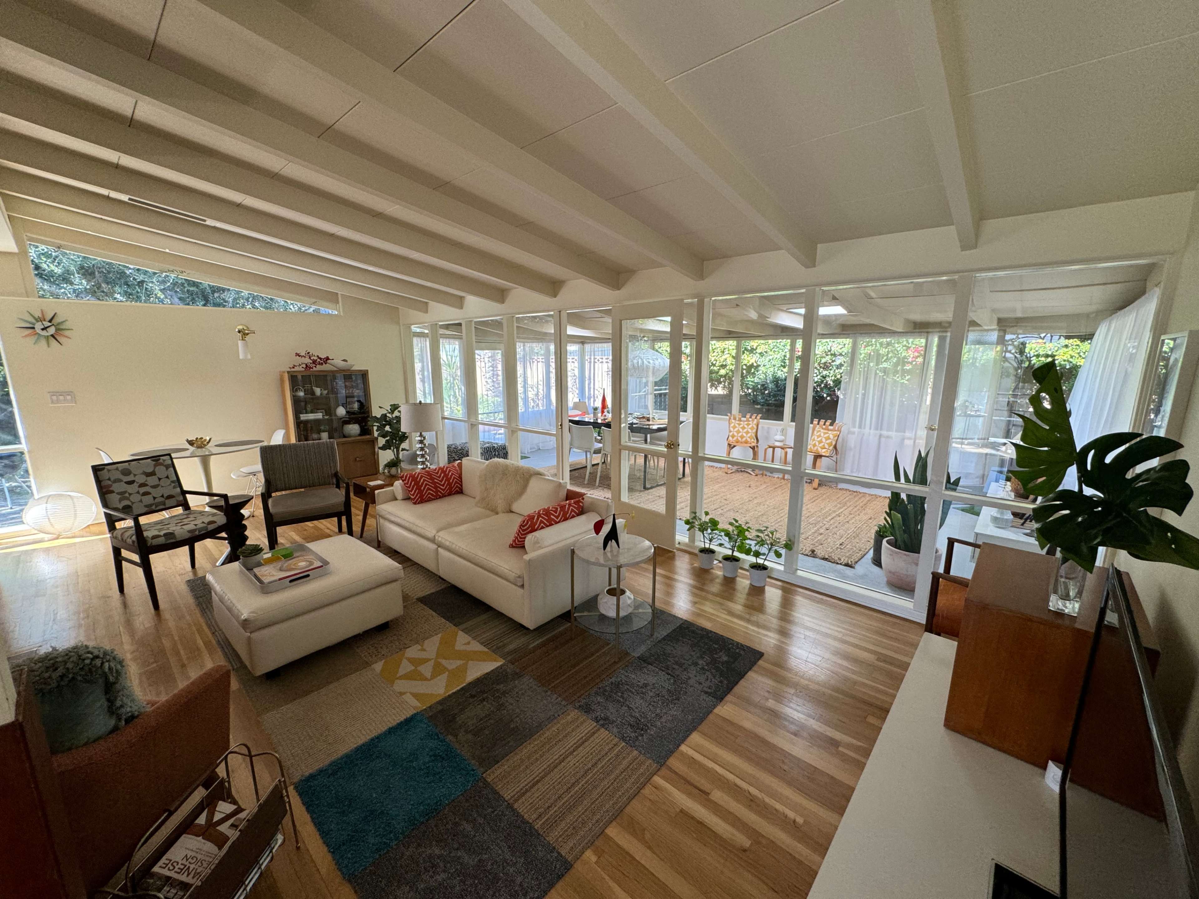 The image shows a bright, airy living room with a white couch, a coffee table, and a view into a sunroom filled with plants and seating.