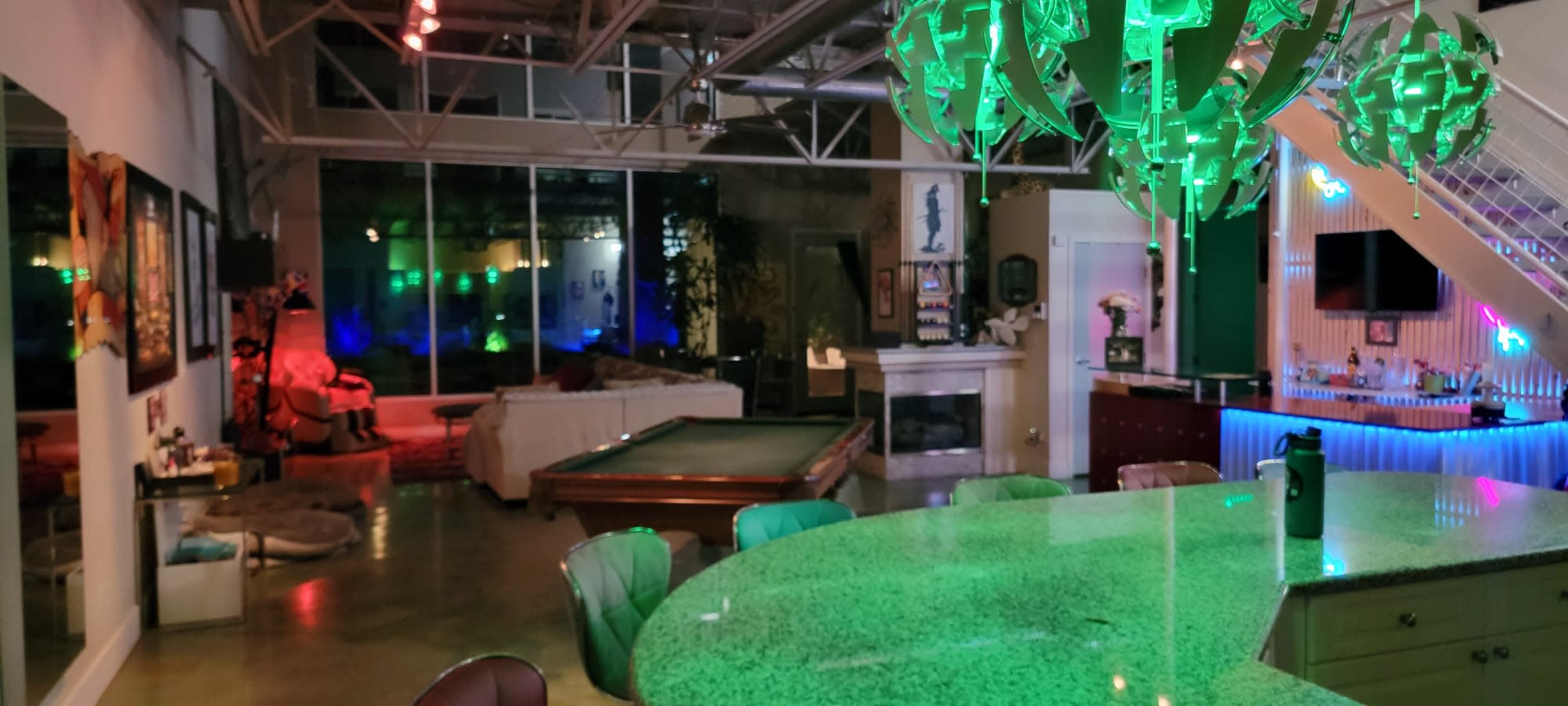A spacious modern loft features a green chandelier, a pool table, and an illuminated bar area, with seating and artwork along the walls.