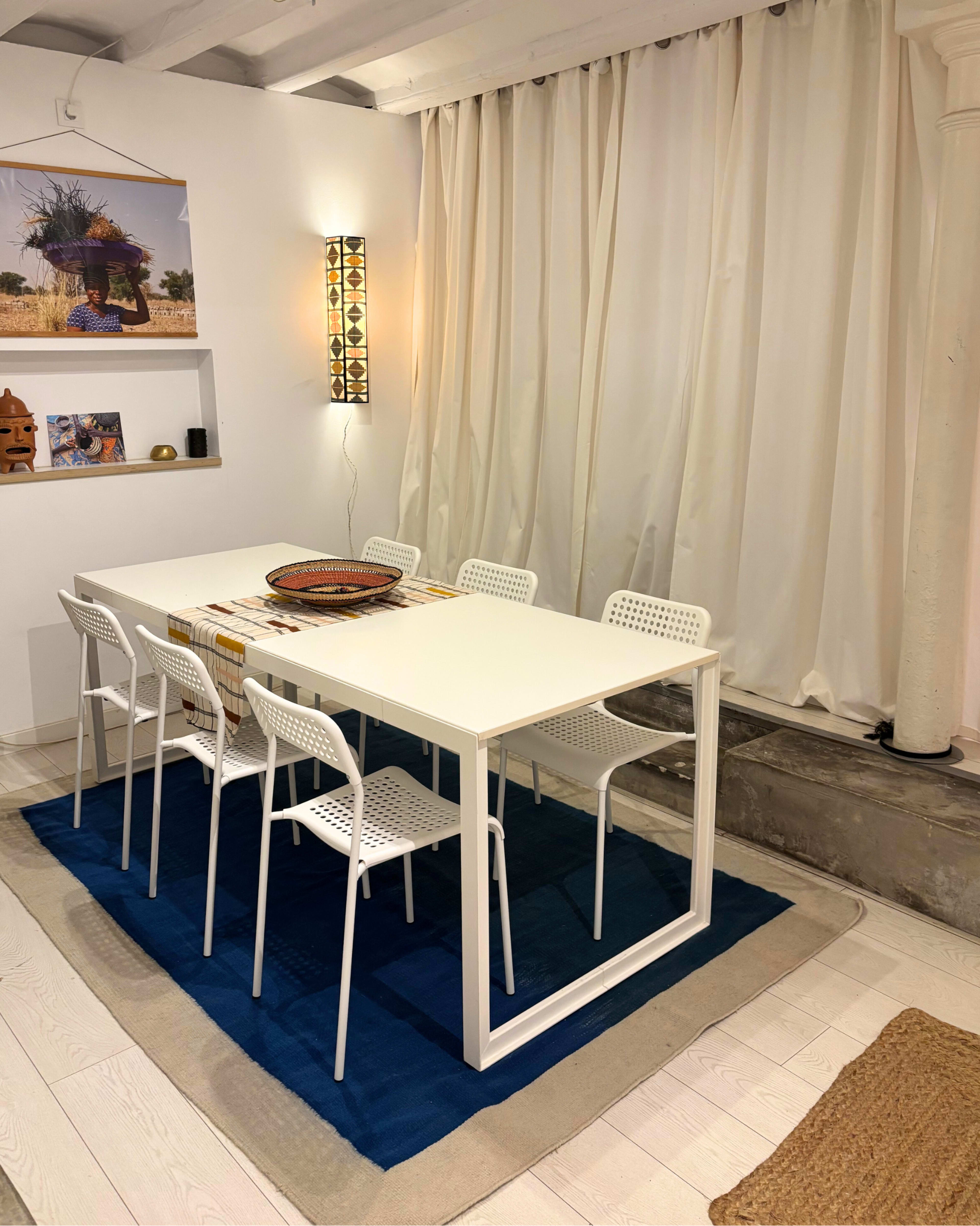 A modern dining area features a white table with six chairs arranged on a blue rug, surrounded by cream-colored curtains and decorative wall art.