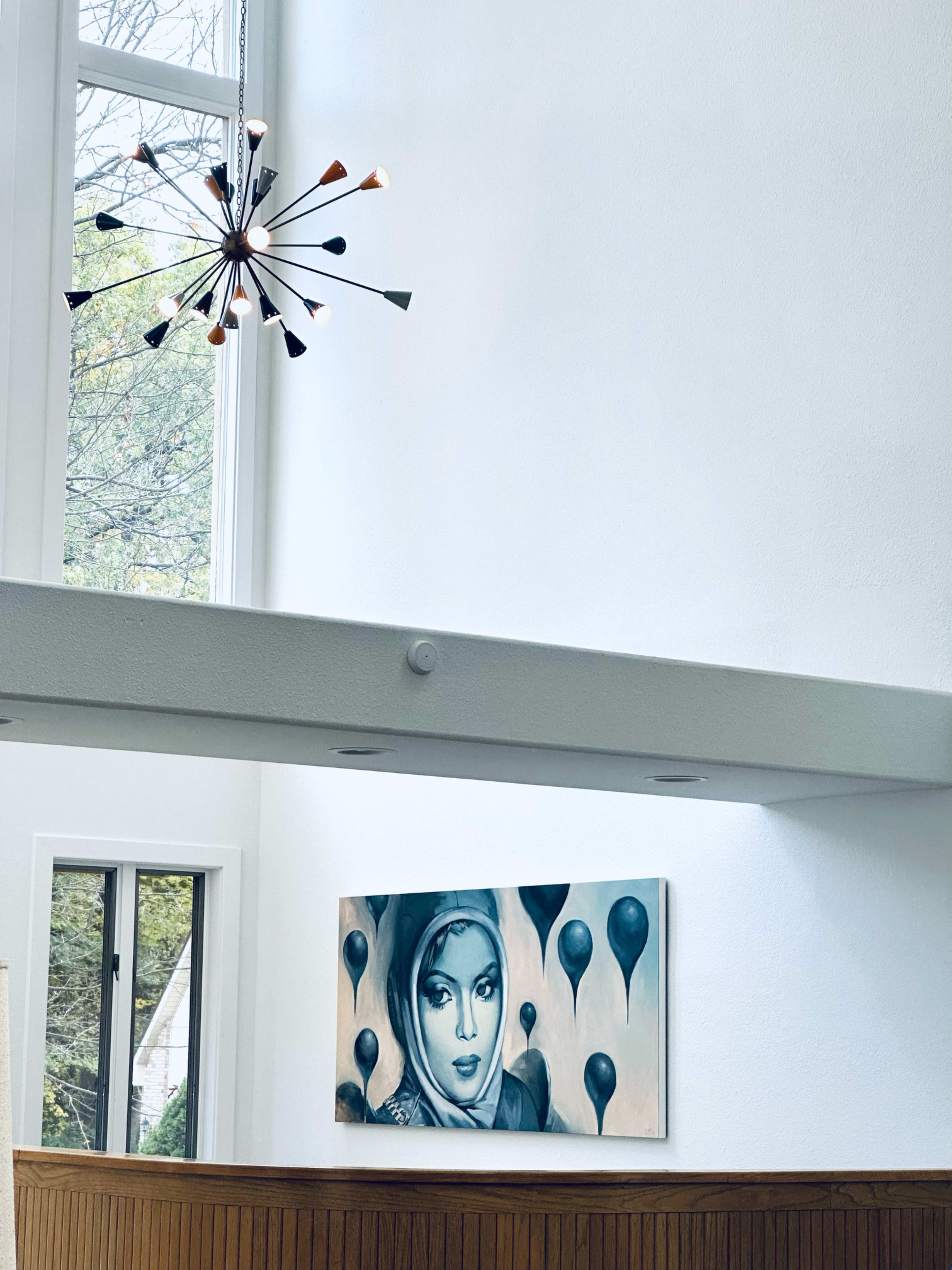 The image shows a modern interior with a large painting of a woman surrounded by blue balloons, illuminated by a unique light fixture above.