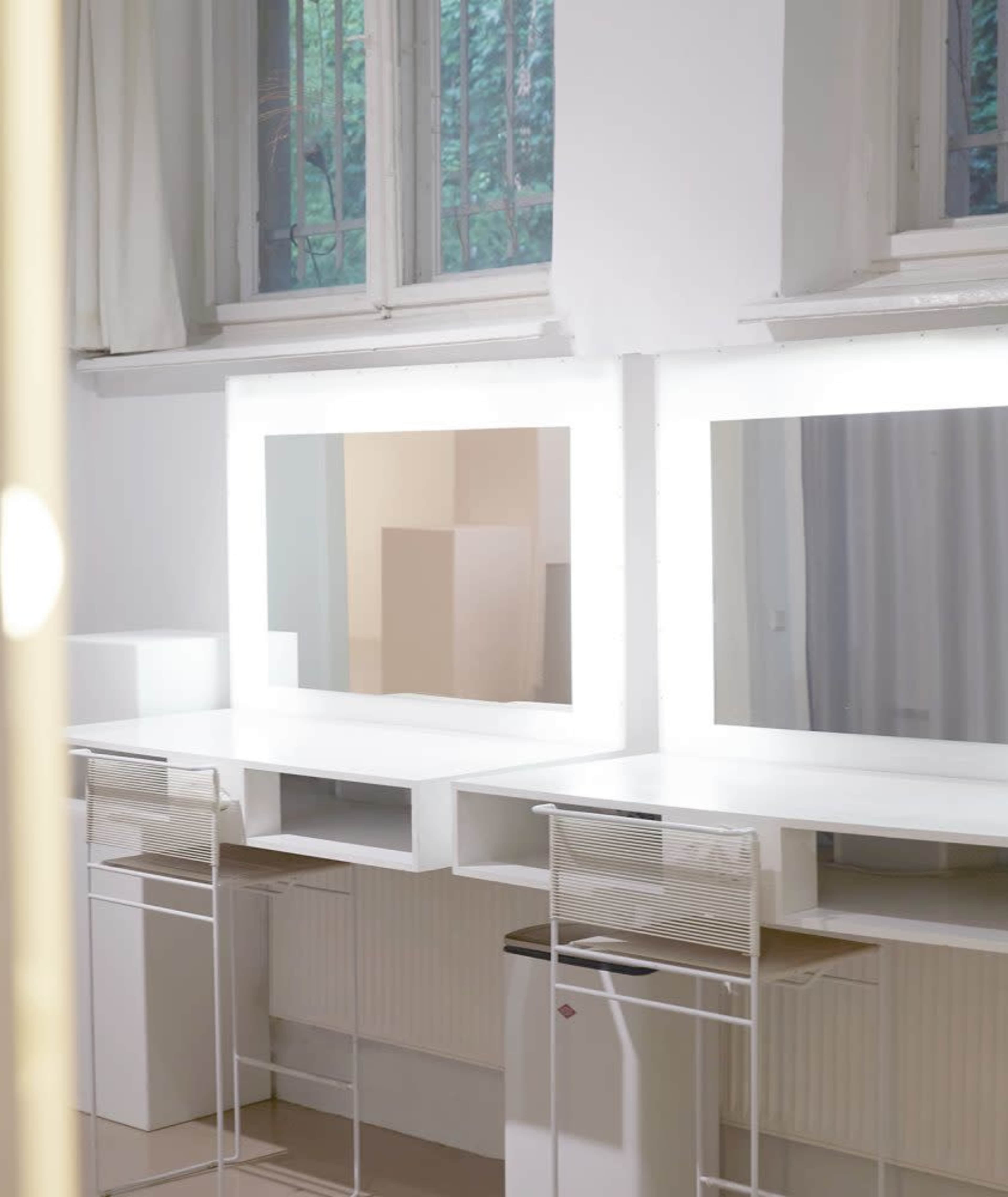 The image shows a bright, minimalist workspace with two large mirrors, white desks, and transparent chairs, set against large windows.