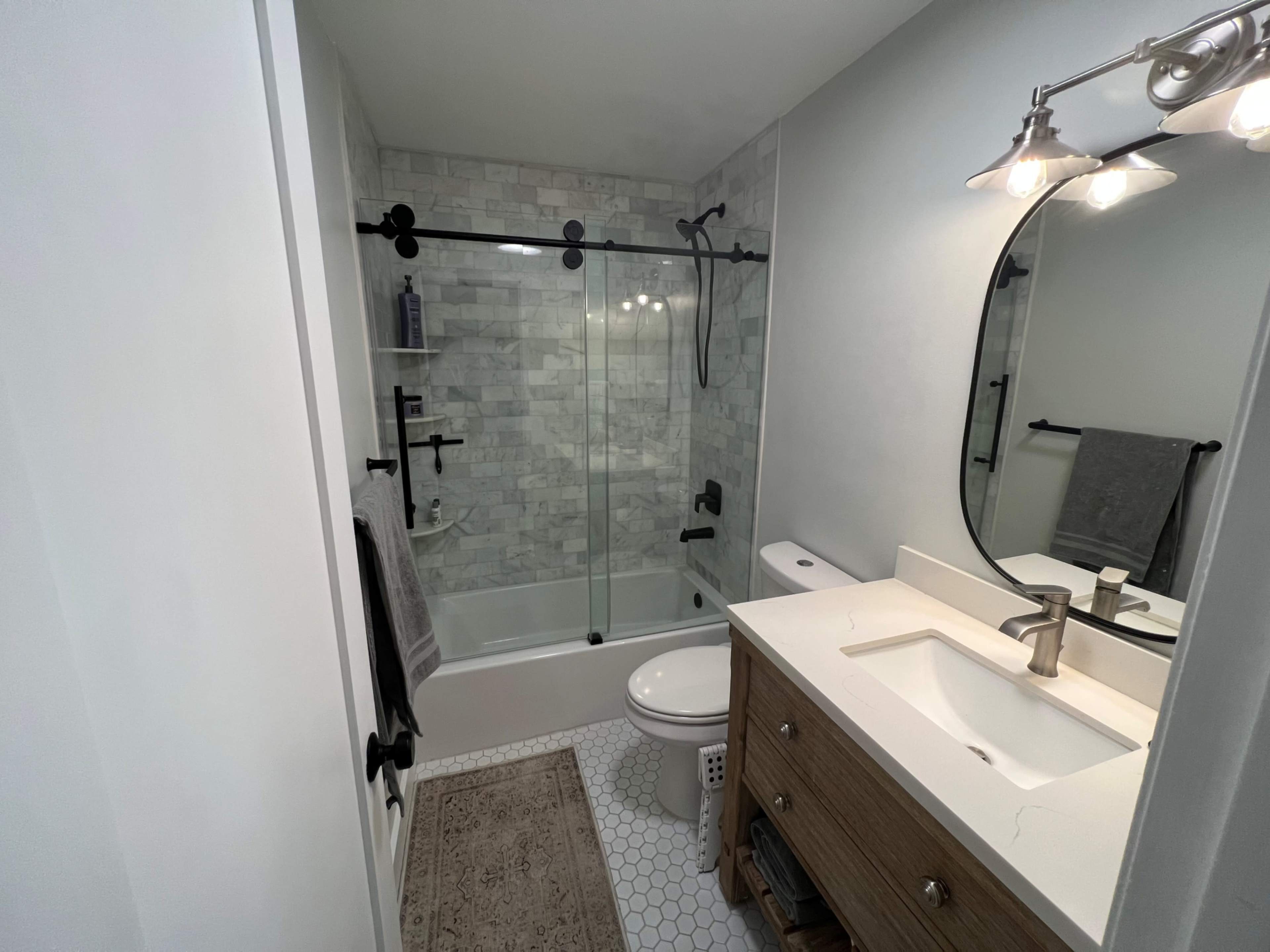 The image shows a modern bathroom featuring a glass shower enclosure, a toilet, a sink with a wooden vanity, and tiled walls.