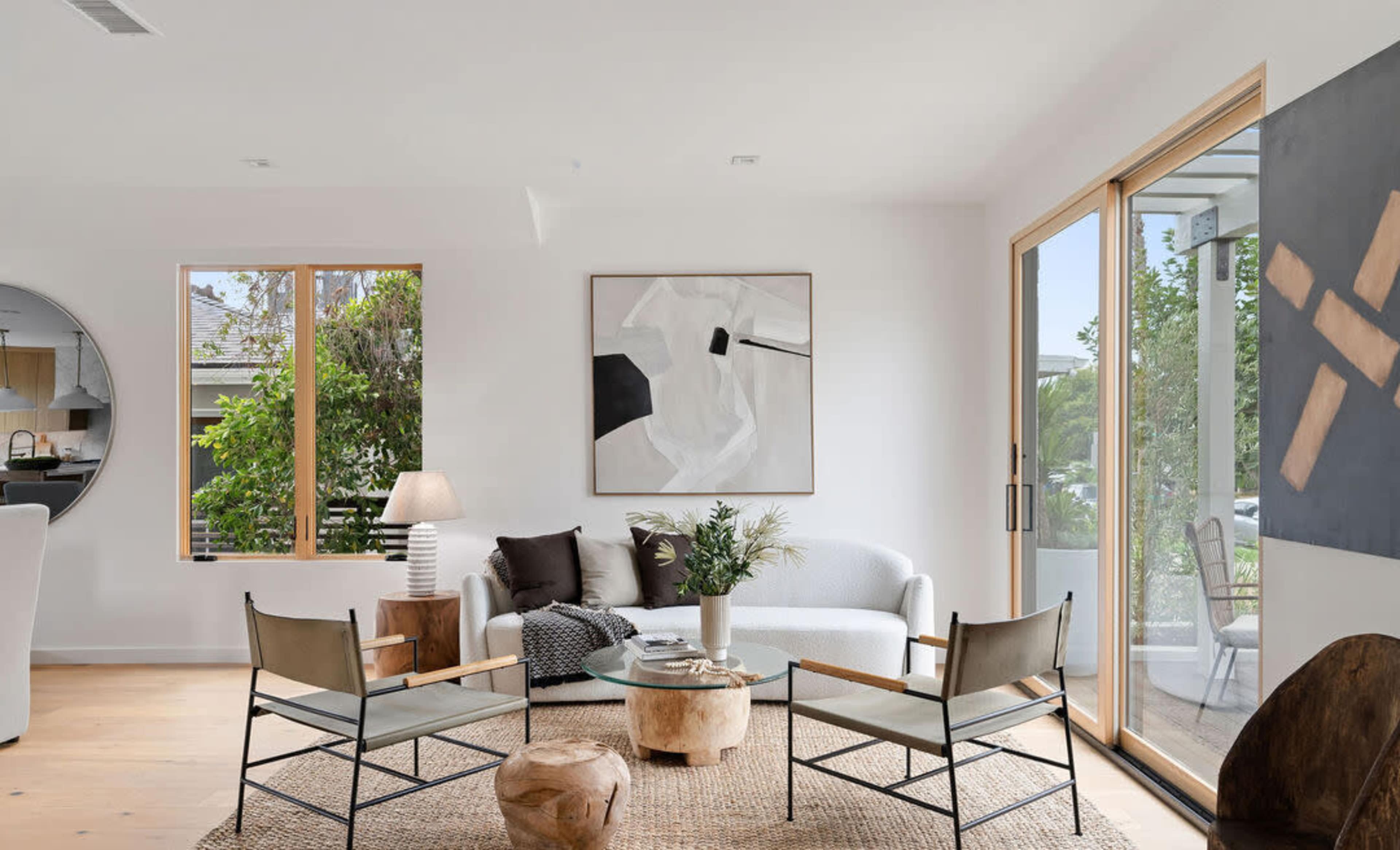 A modern living room features a light gray sofa, two metal-framed chairs, a round side table, and large windows that let in natural light.