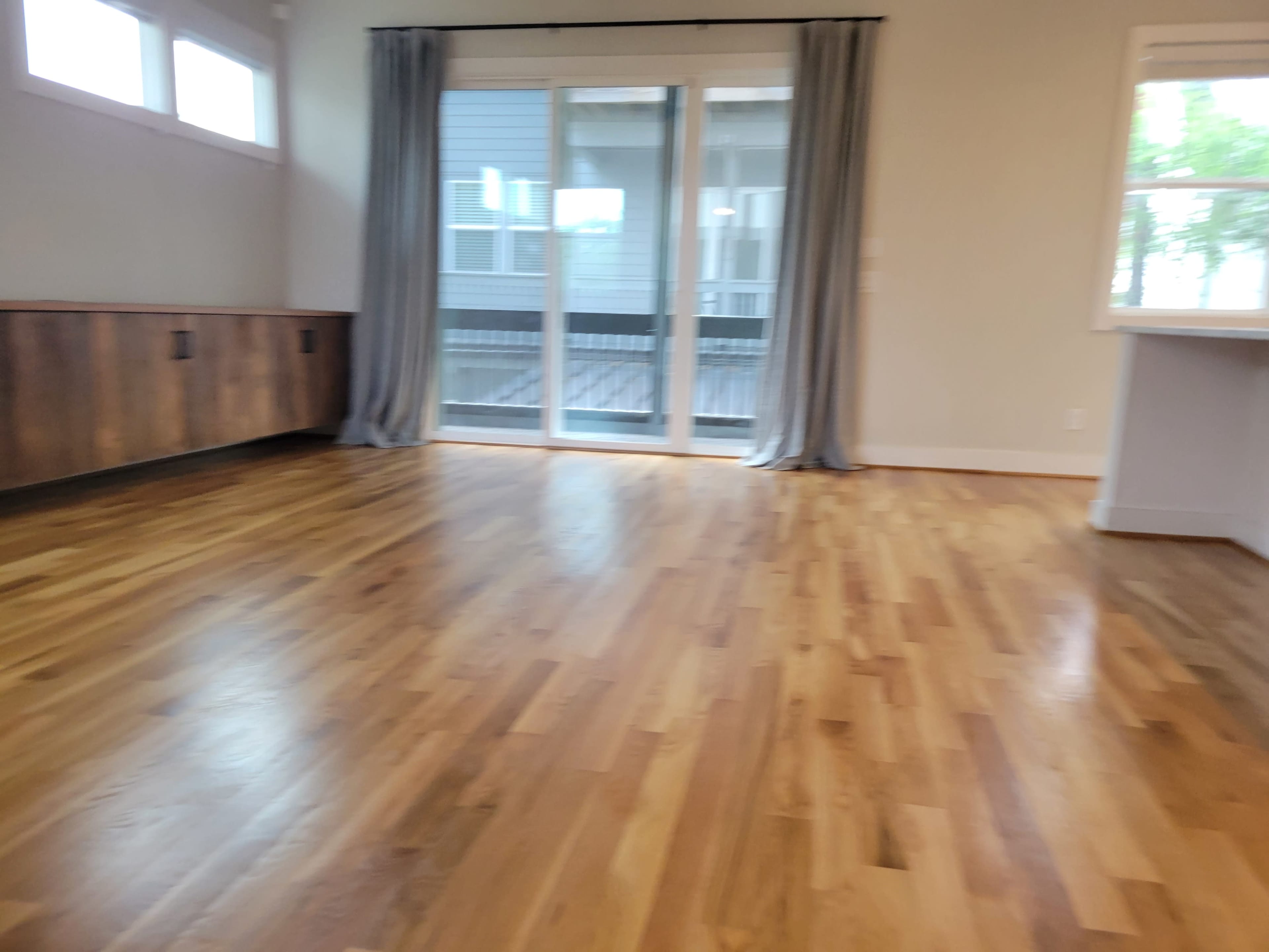 The image shows a spacious room with wooden floors, large sliding doors that lead to a balcony, and gray curtains on either side.