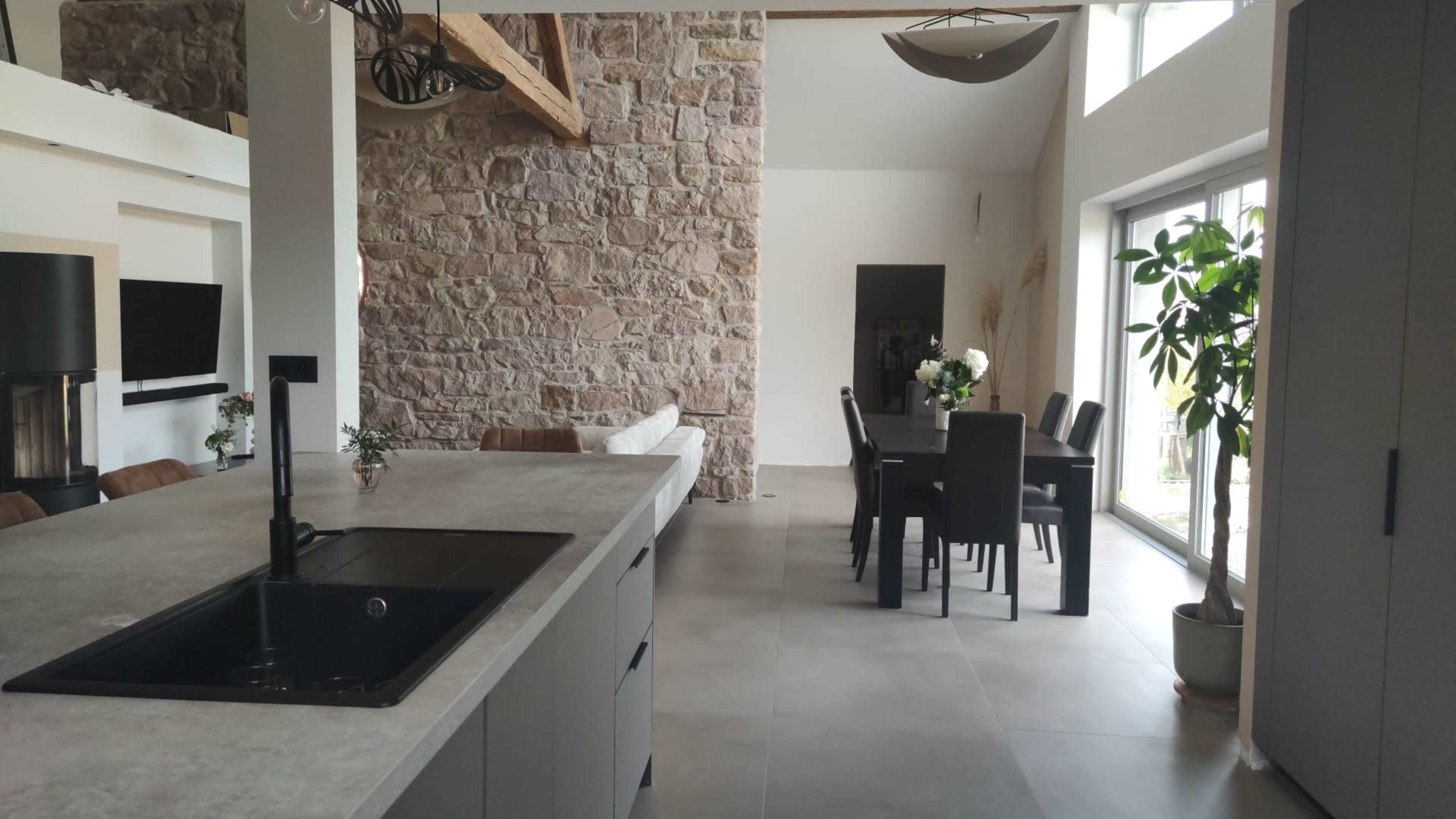 The image shows a modern kitchen with an island, featuring a stone wall in the background and a dining area with a table and chairs to the right.