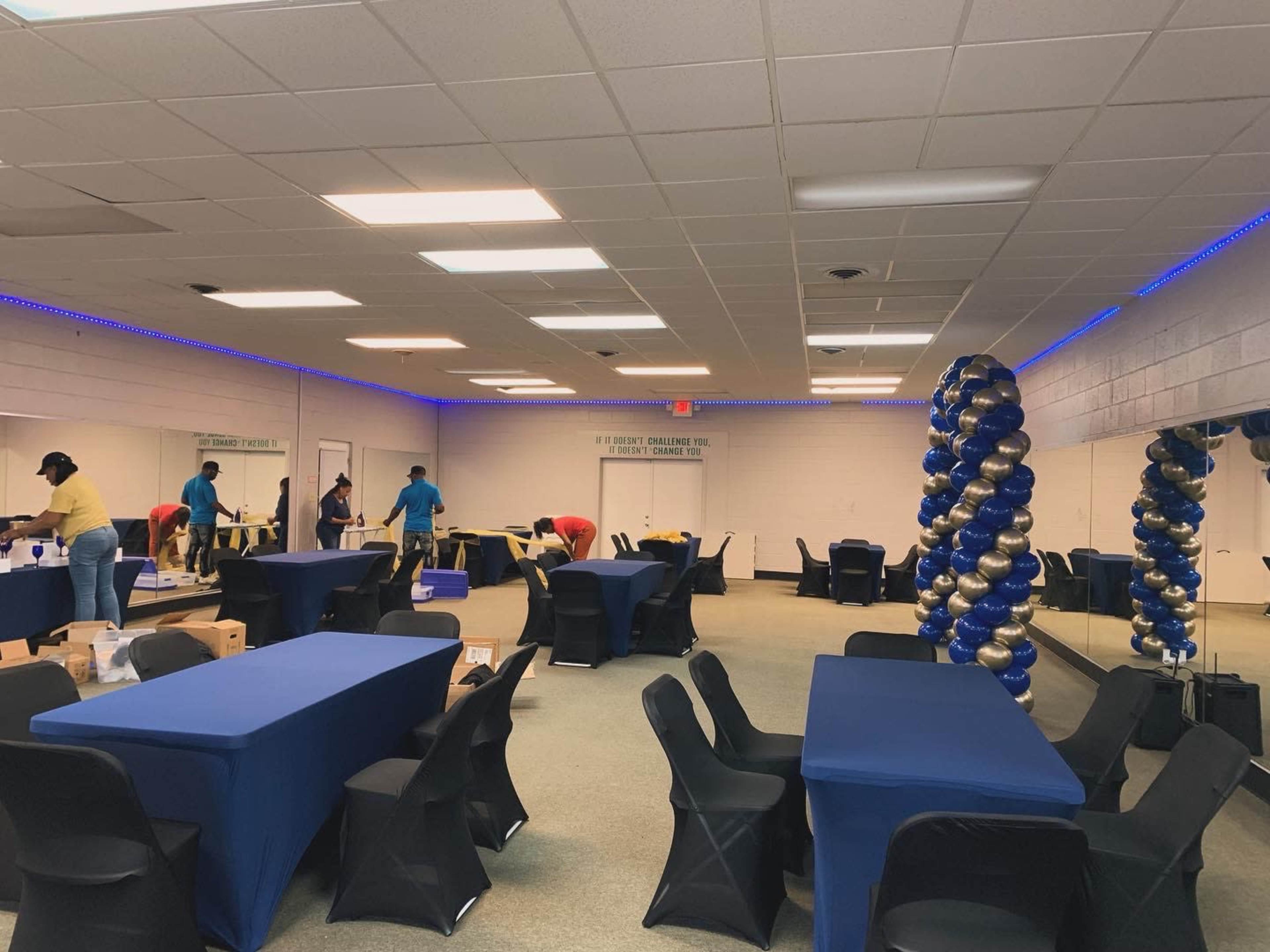 A group of people is setting up tables and chairs in a large room decorated with blue and gold balloons.