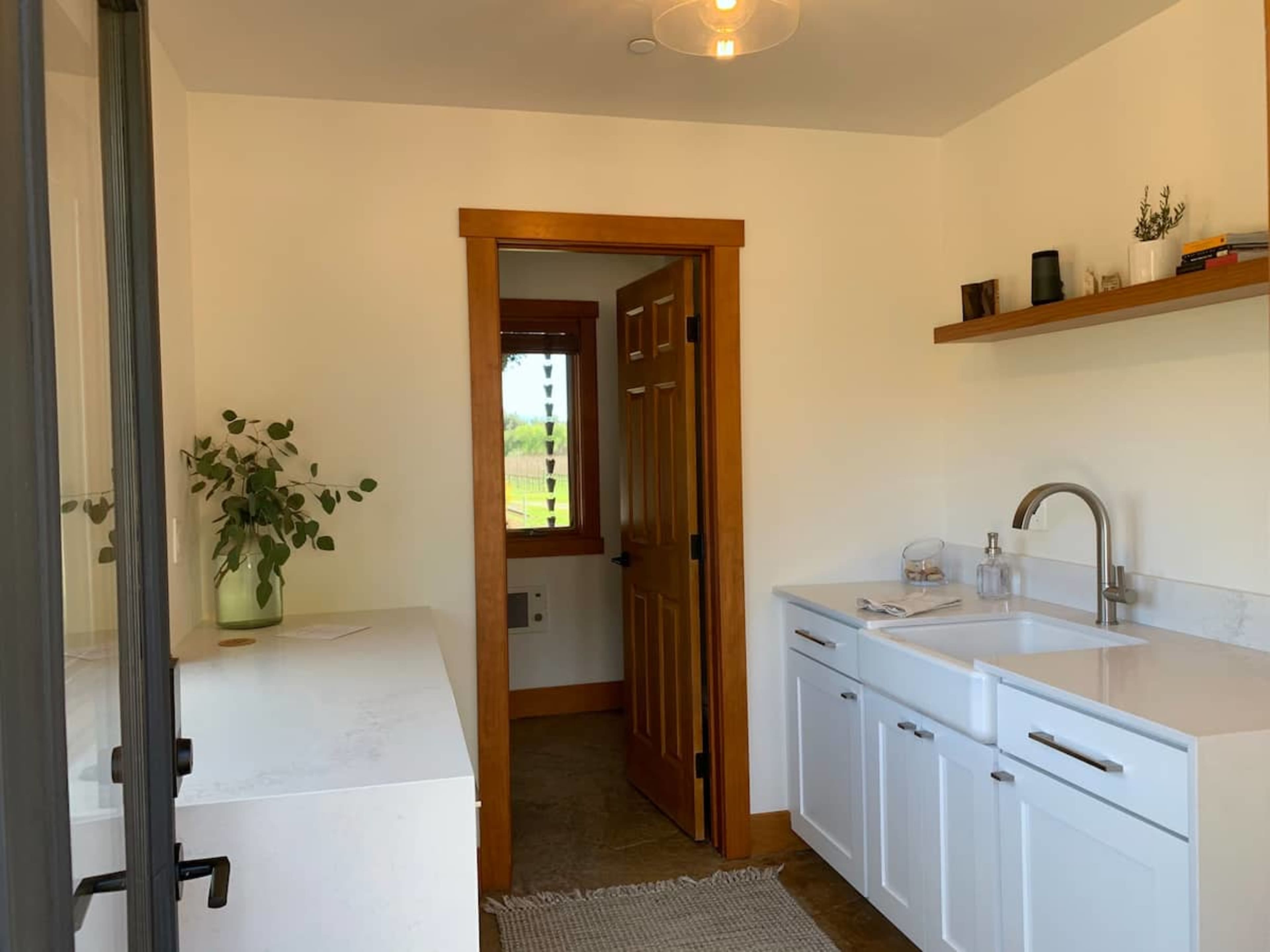 The image shows a modern bathroom with a white sink and cabinet, a door leading to another room, and a shelf with decorative items.