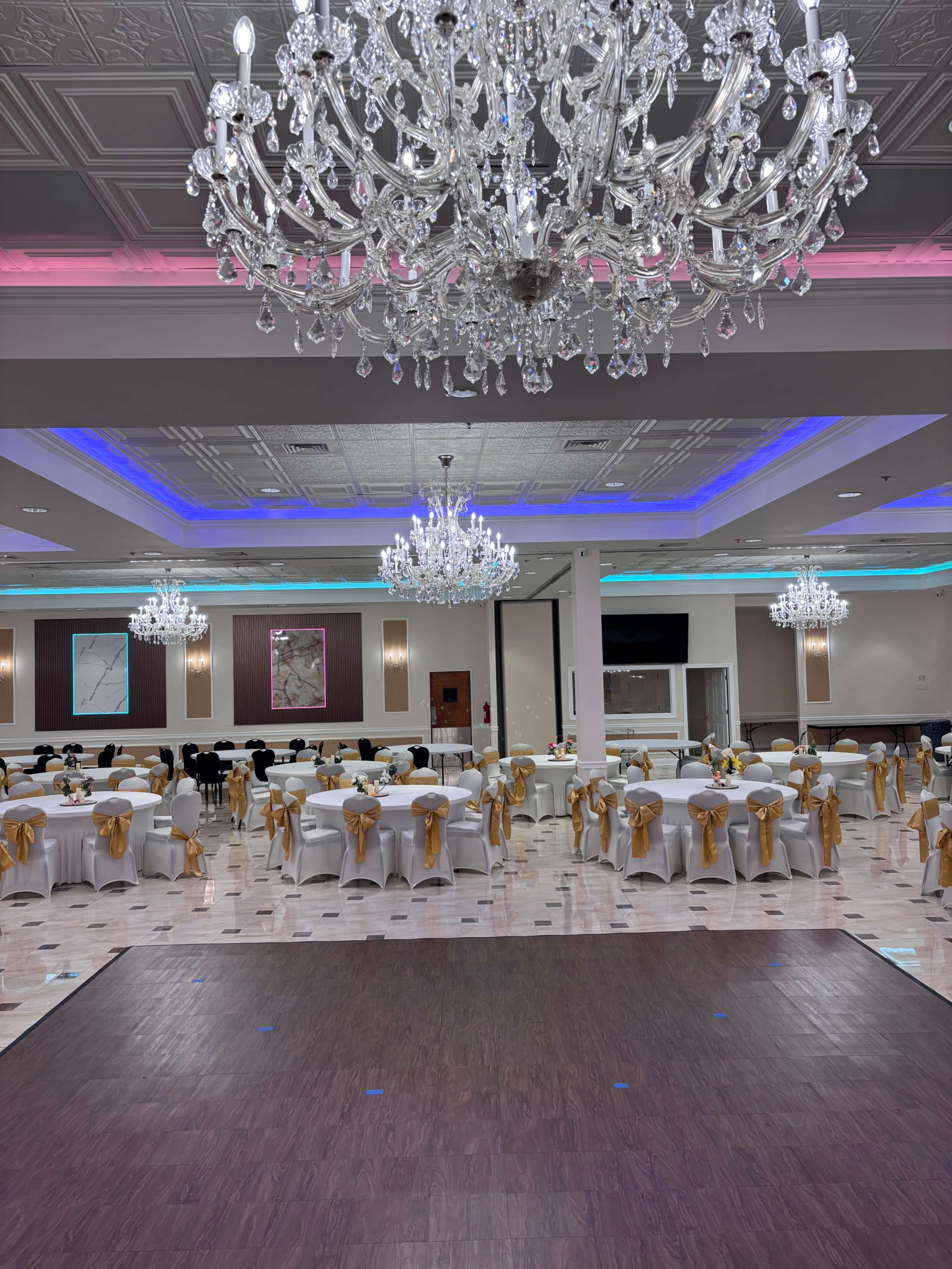 The image shows a banquet hall with elegant chandeliers, tables covered in white tablecloths with yellow sashes, and colorful lighting along the ceiling.