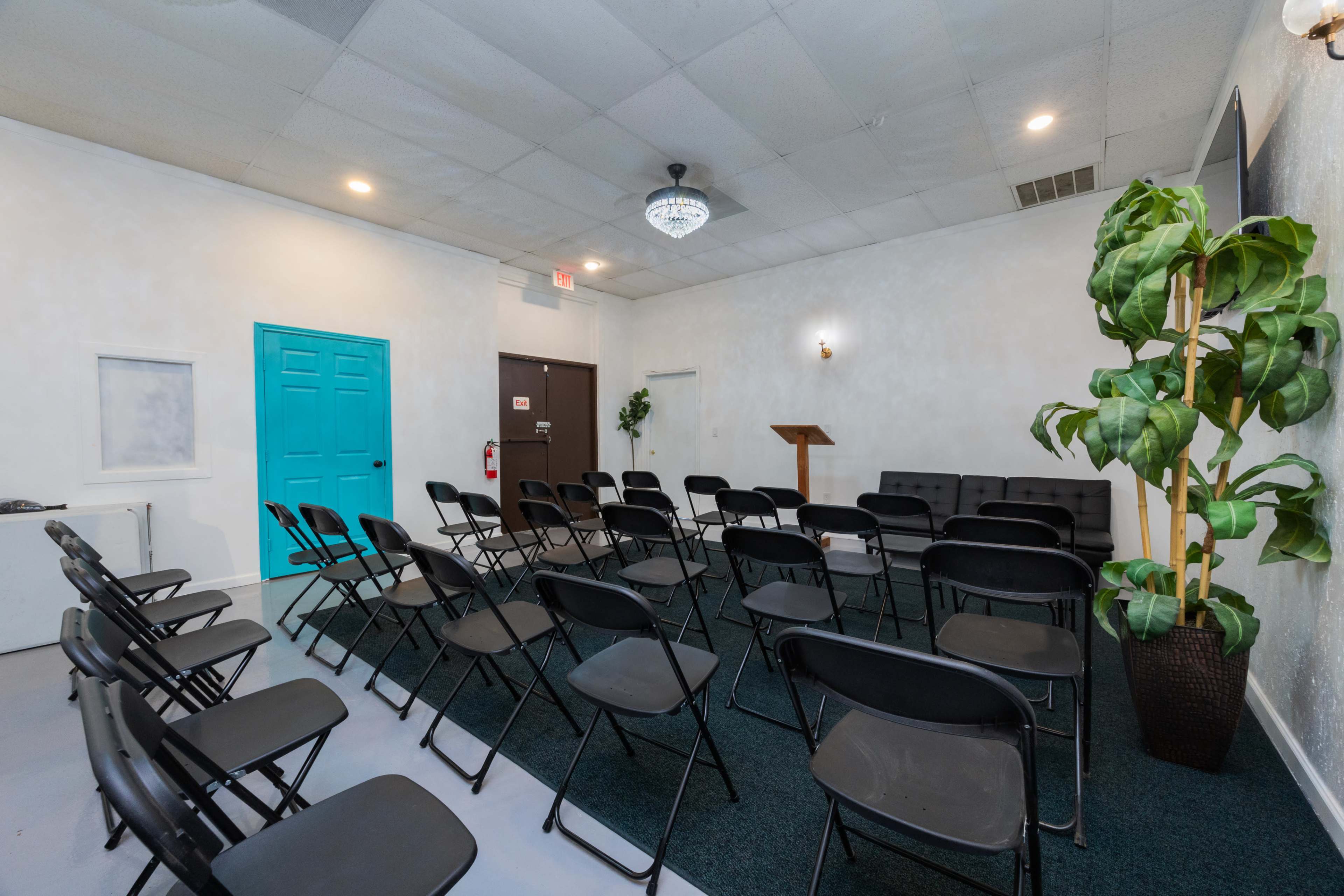 A room set up with black folding chairs arranged in rows facing a small podium, with a blue door and a large plant in a pot visible on one side.
