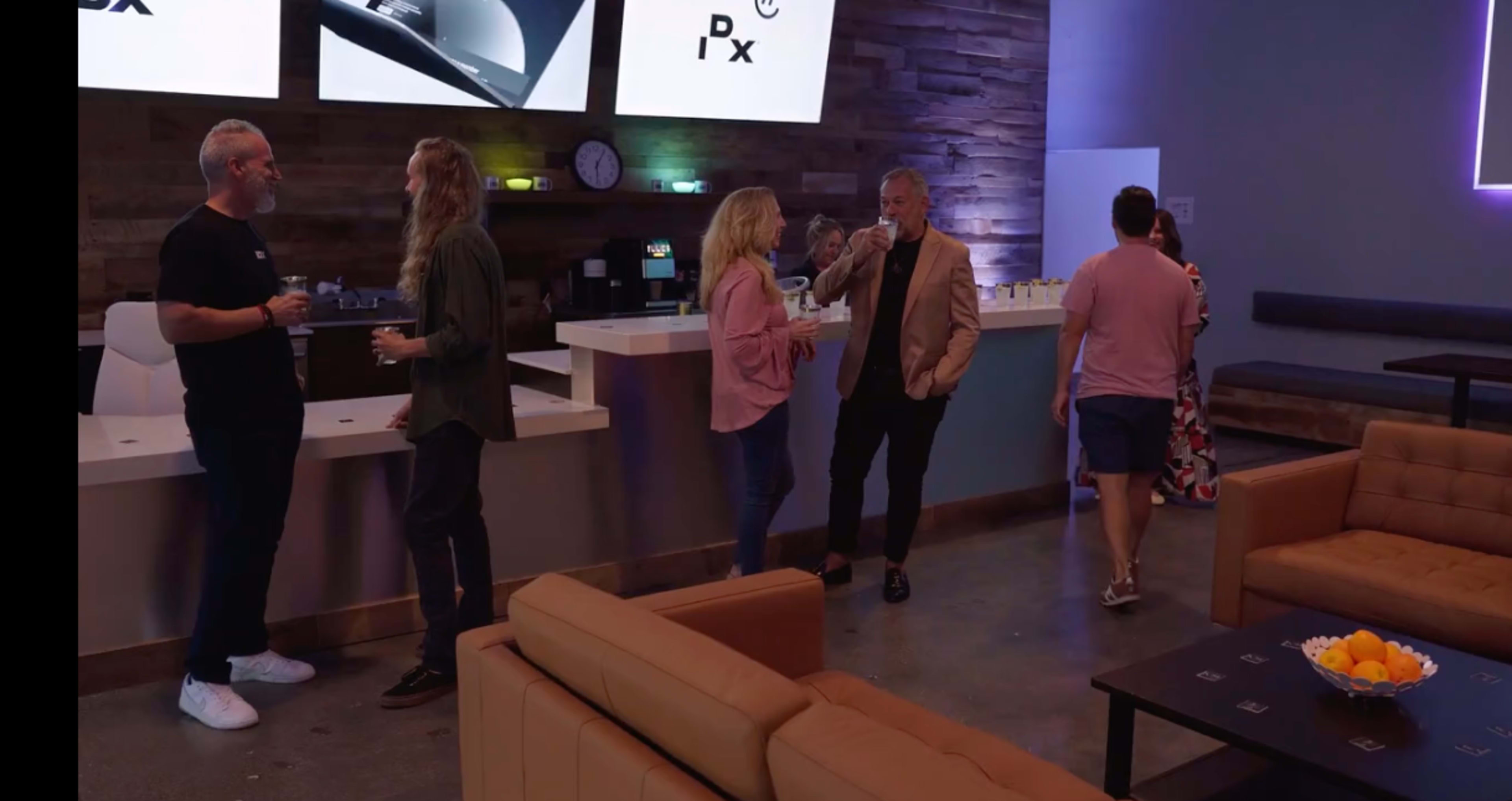 A group of people stands in a modern lounge area with a bar, chatting and holding drinks, while a large screen displays the logo "Pix."