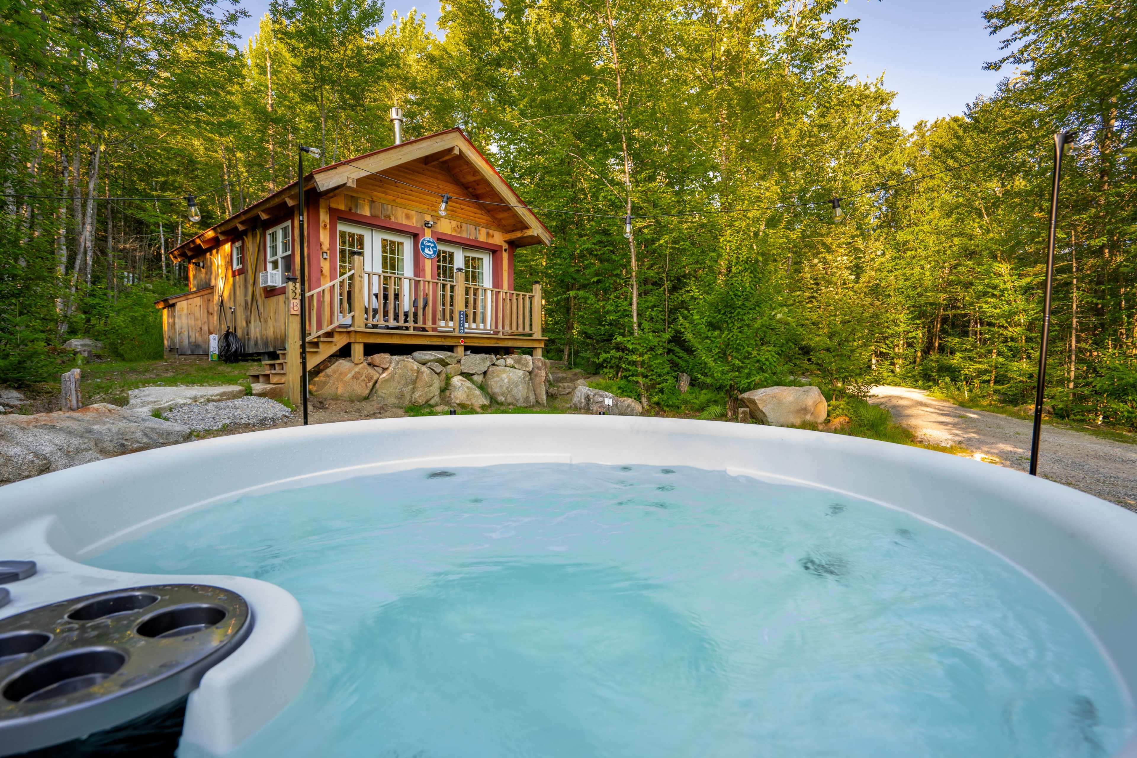 A wooden cabin with a porch is surrounded by trees, and a hot tub filled with water is positioned in the foreground.