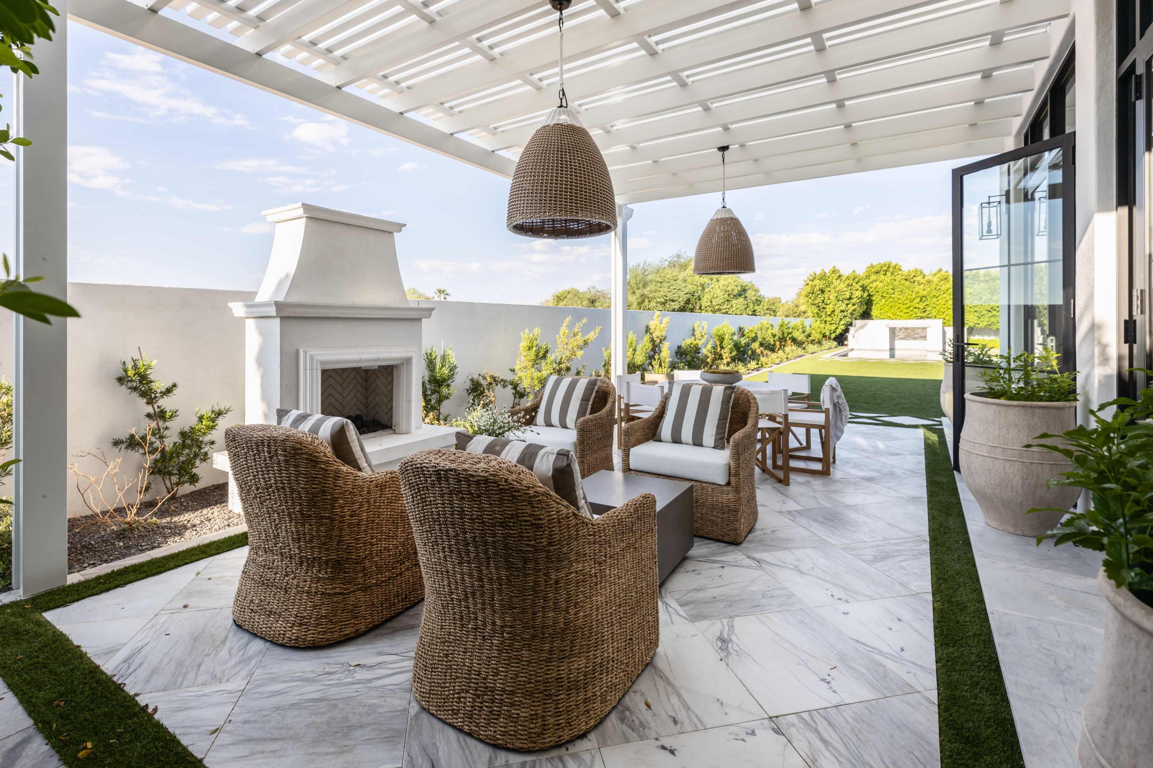 The image shows a modern patio area featuring wicker chairs, a sofa with striped cushions, a fireplace, and a well-maintained lawn in the background.