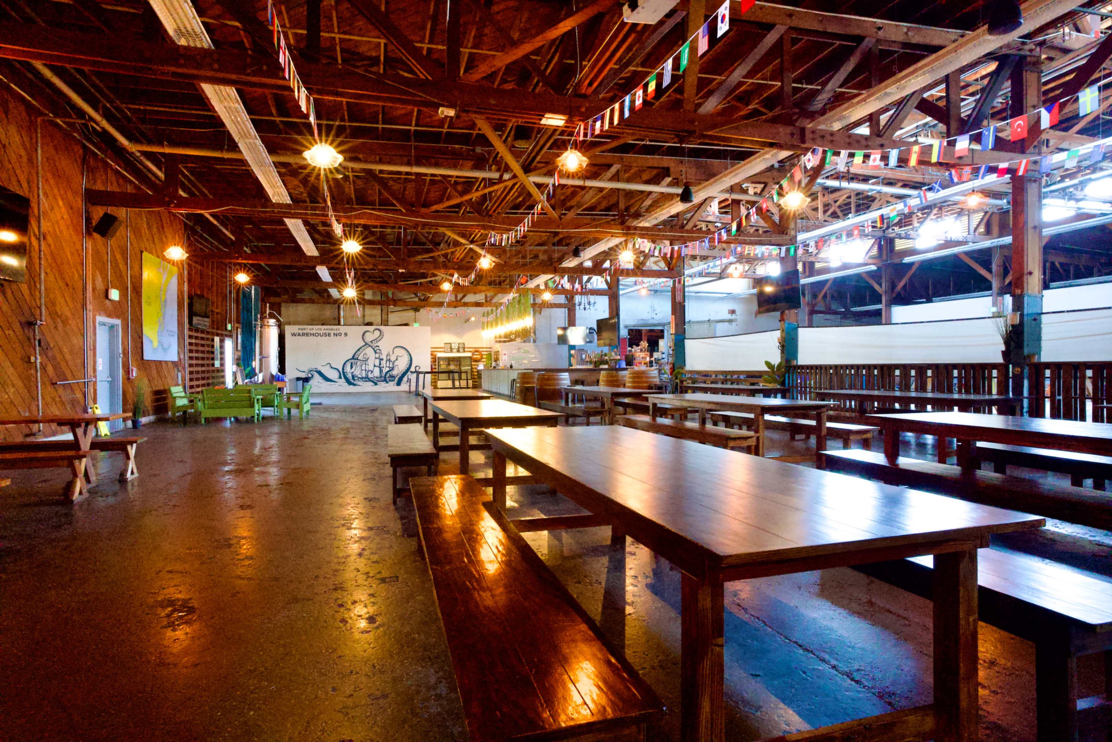 The image shows a spacious, industrial-style dining hall with wooden tables, benches, and colorful flags hanging from the ceiling.