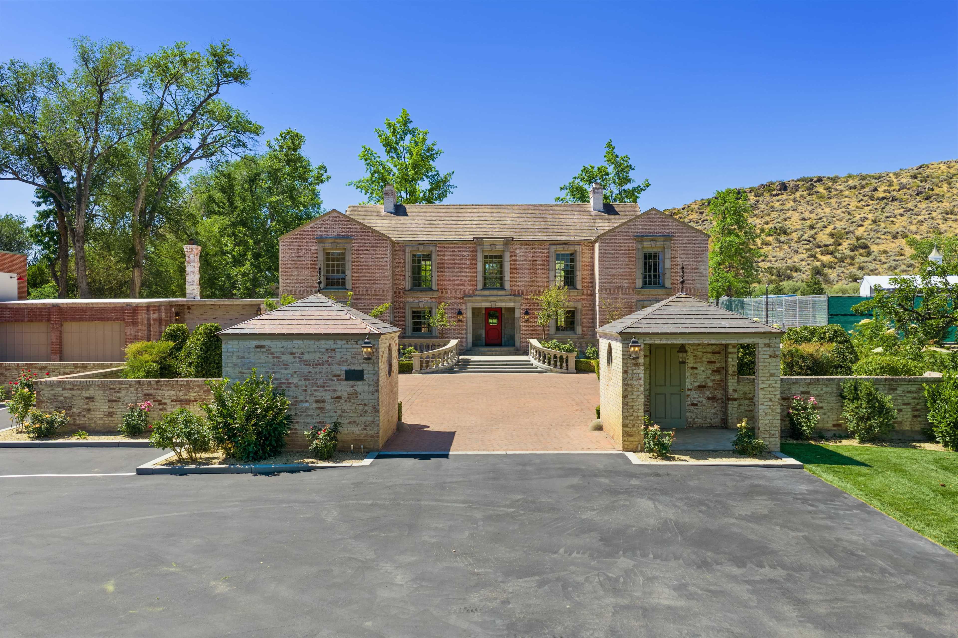 The image shows a large brick house with a manicured front yard and a paved driveway, flanked by trees and shrubs against a clear blue sky.