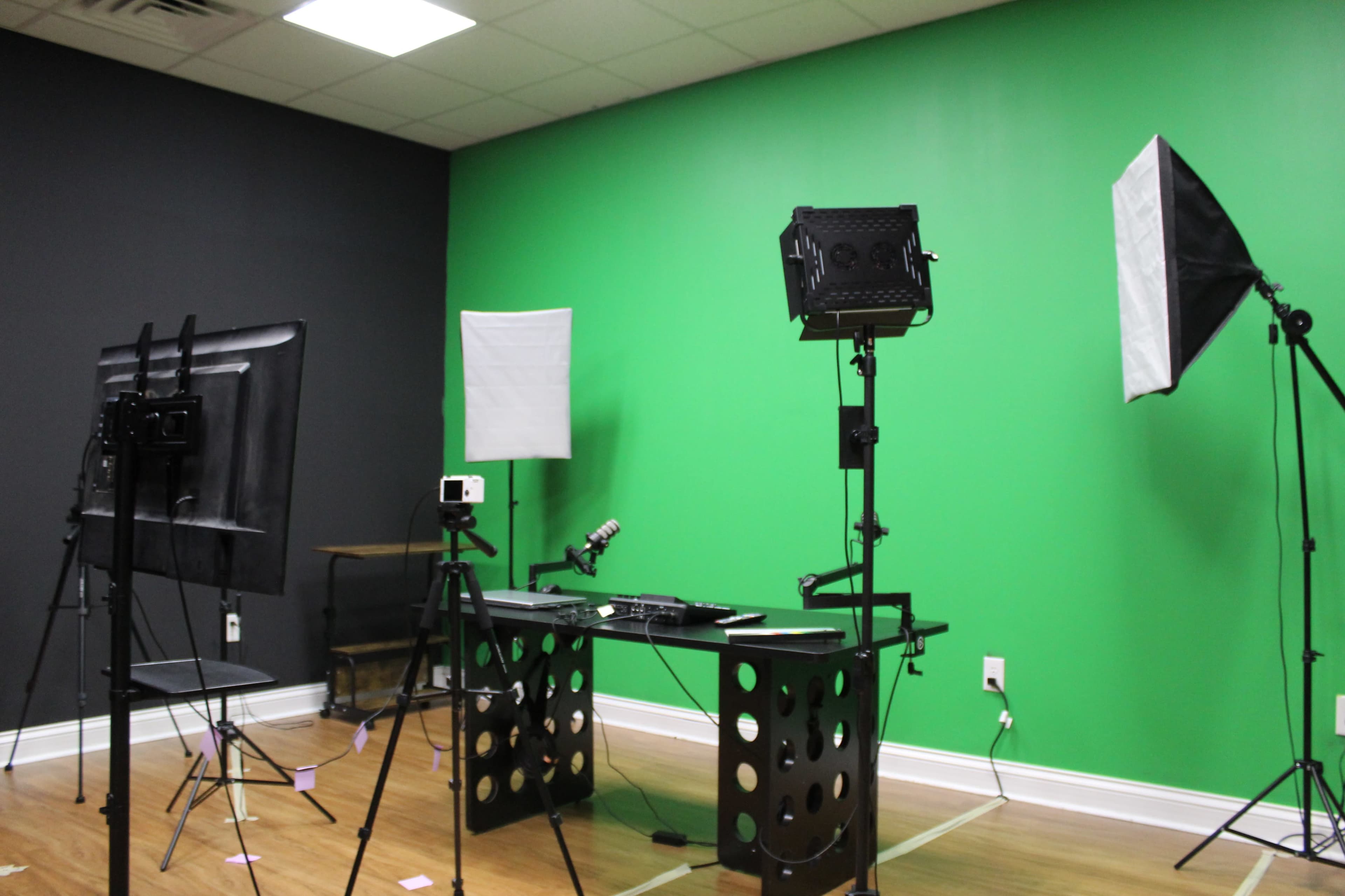 The image shows a studio setup with a green wall, two lighting stands, a microphone, a camera on a tripod, and a table with equipment.